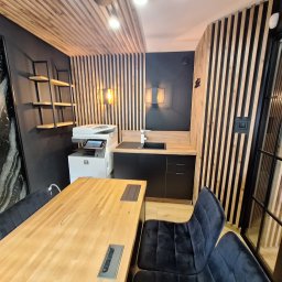 Od remontu do efektu - remonty i wykończenia wnętrz - Nowoczesne biuro z drewnianymi lamelami na ścianach i suficie, czarnymi meblami kuchennymi i dużym stołem z krzesłami. Widoczny ekspres do kawy i dekoracyjny panel.