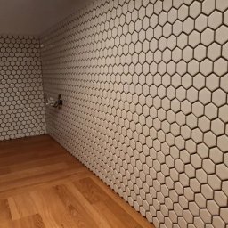 Od remontu do efektu - remonty i wykończenia wnętrz - Wykończona łazienka z heksagonalnymi, białymi płytkami na ścianach i drewnianą podłogą. Widoczny fragment instalacji wodnej.