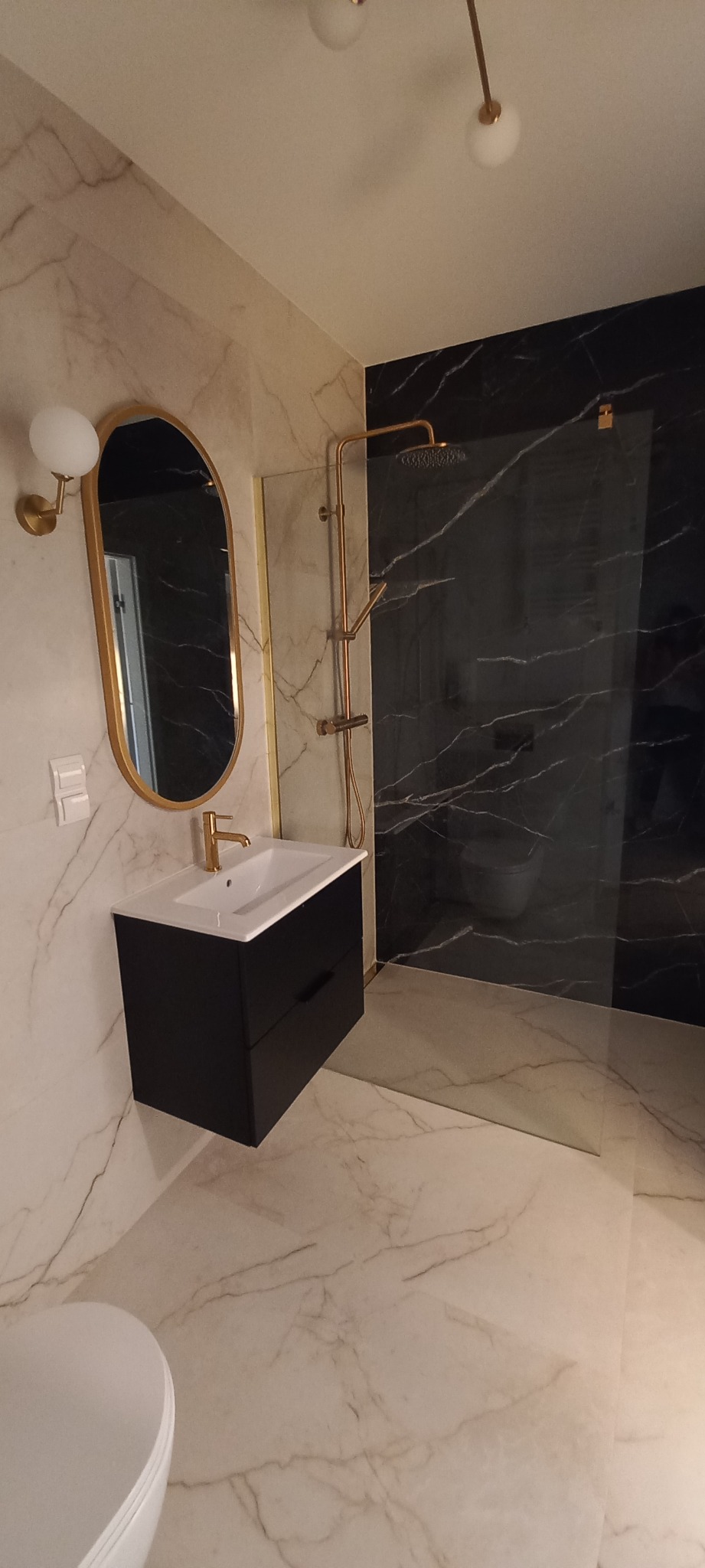 Nowoczesna łazienka z czarną szafką pod umywalką, złotą armaturą i kabiną prysznicową z czarnym marmurem. Całość wykończona płytkami imitującymi biały marmur.