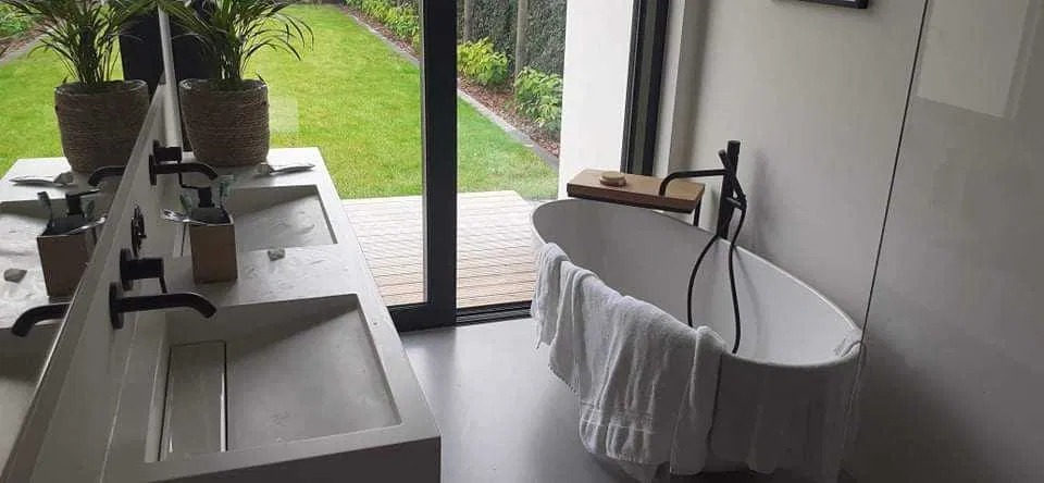 Nowoczesna łazienka z wolnostojącą wanną, podwójną umywalką z czarną armaturą i dużym oknem z widokiem na ogród. Minimalistyczny design, jasne kolory, rośliny doniczkowe.