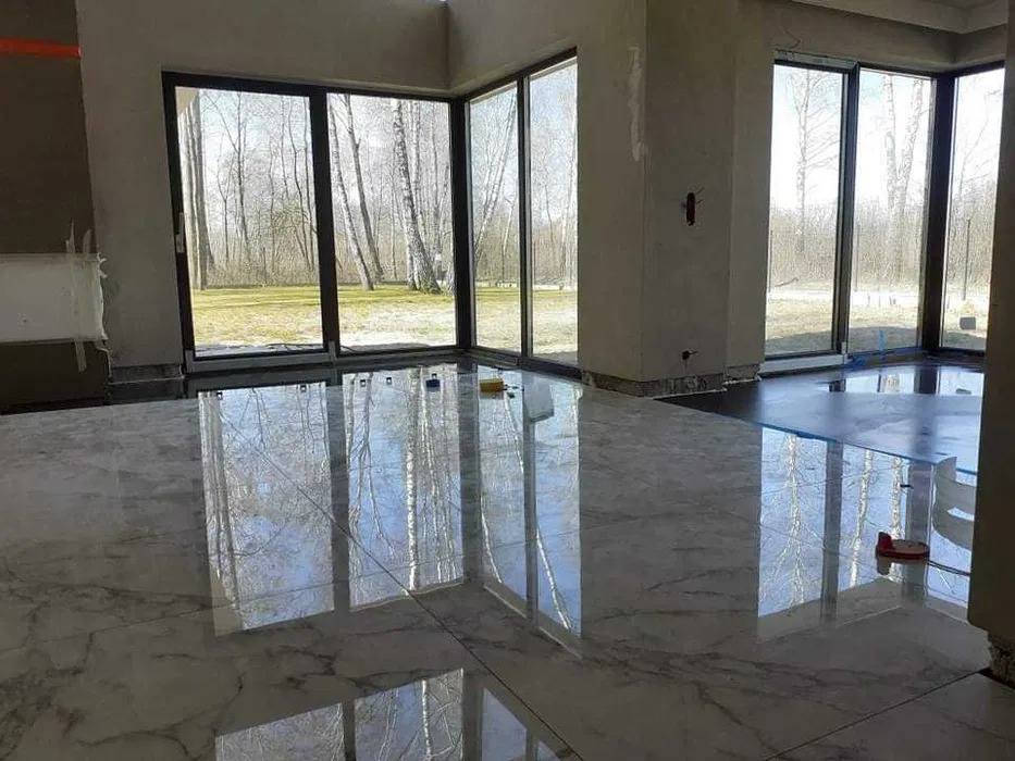 Błyszcząca, marmurowa podłoga w jasnym wnętrzu z dużymi oknami, przez które widać las. Częściowo wykończone ściany i widoczne narzędzia sugerują etap prac wykończeniowych.