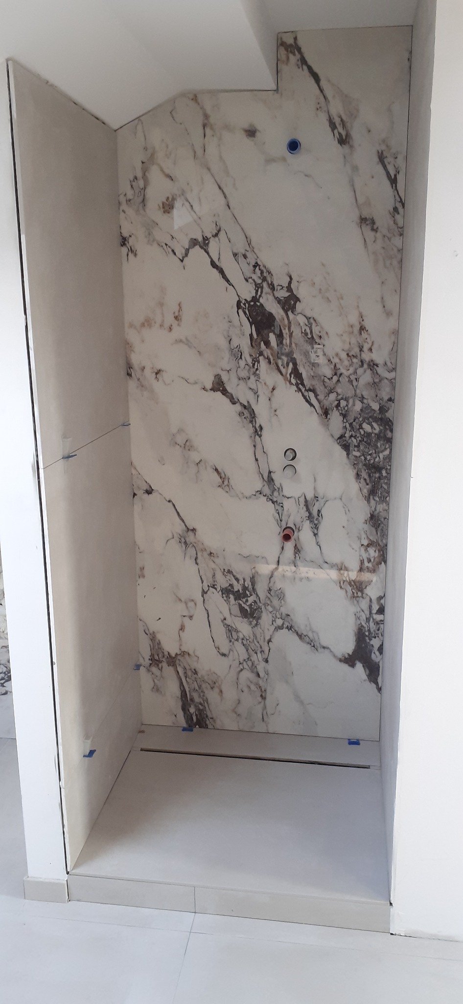 Wykończona wnęka prysznicowa z marmurową płytą na ścianie i odpływem liniowym w podłodze. Widoczne przyłącza wodne. Minimalistyczny design.