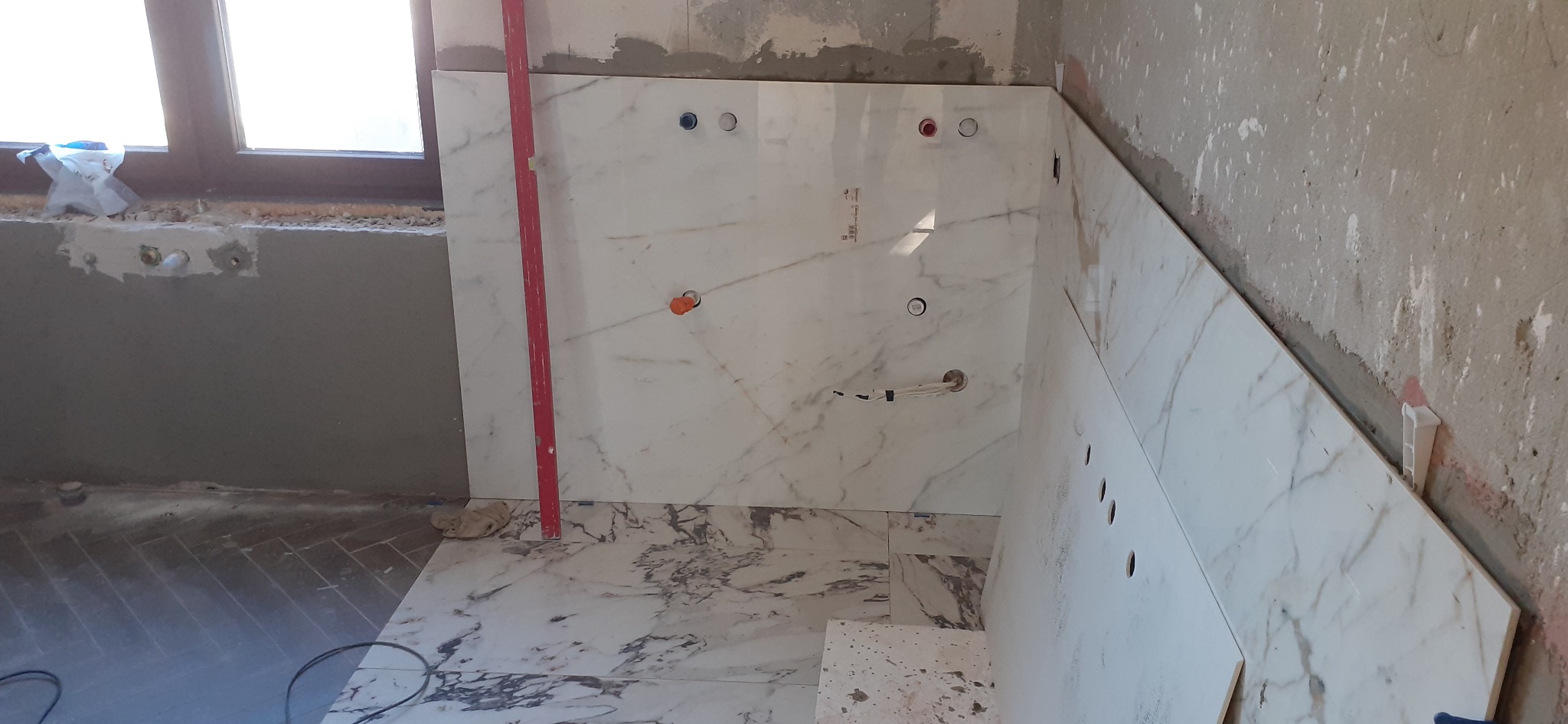 Układanie marmurowych płytek w łazience, widoczne otwory na instalacje, poziomica przy ścianie, surowy stan ścian. Podłoga częściowo wyłożona płytkami w jodełkę.