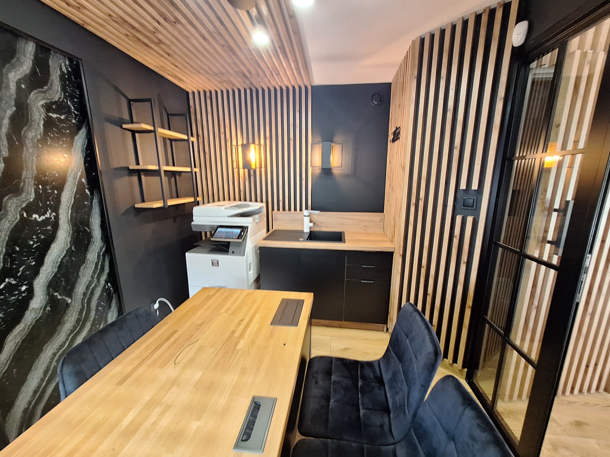 Nowoczesne biuro z drewnianymi lamelami na ścianach i suficie, czarnymi meblami kuchennymi i dużym stołem z krzesłami. Widoczny ekspres do kawy i dekoracyjny panel.