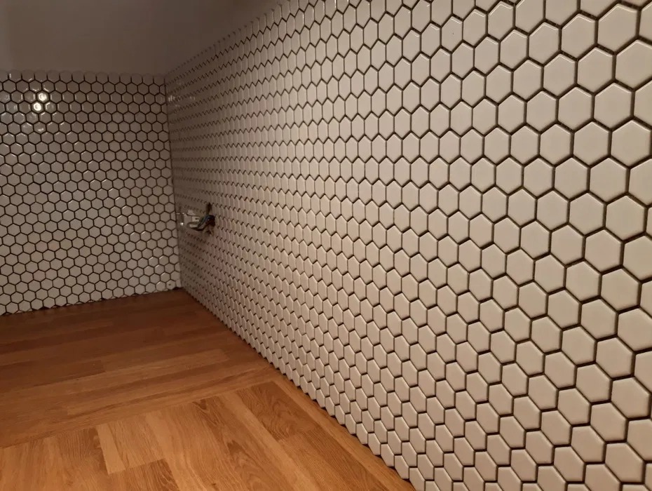 Wykończona łazienka z heksagonalnymi, białymi płytkami na ścianach i drewnianą podłogą. Widoczny fragment instalacji wodnej.