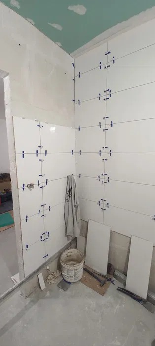 Białe płytki z niebieskimi krzyżykami dystansowymi na ścianie łazienki w trakcie układania. Widoczne narzędzia, wiadro i płyty. Ściana przygotowana pod płytki.