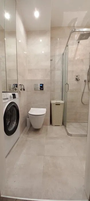 Jasna łazienka po remoncie: kabina prysznicowa, toaleta podwieszana, pralka i kosz na pranie. Płytki w odcieniach beżu, minimalistyczny styl.