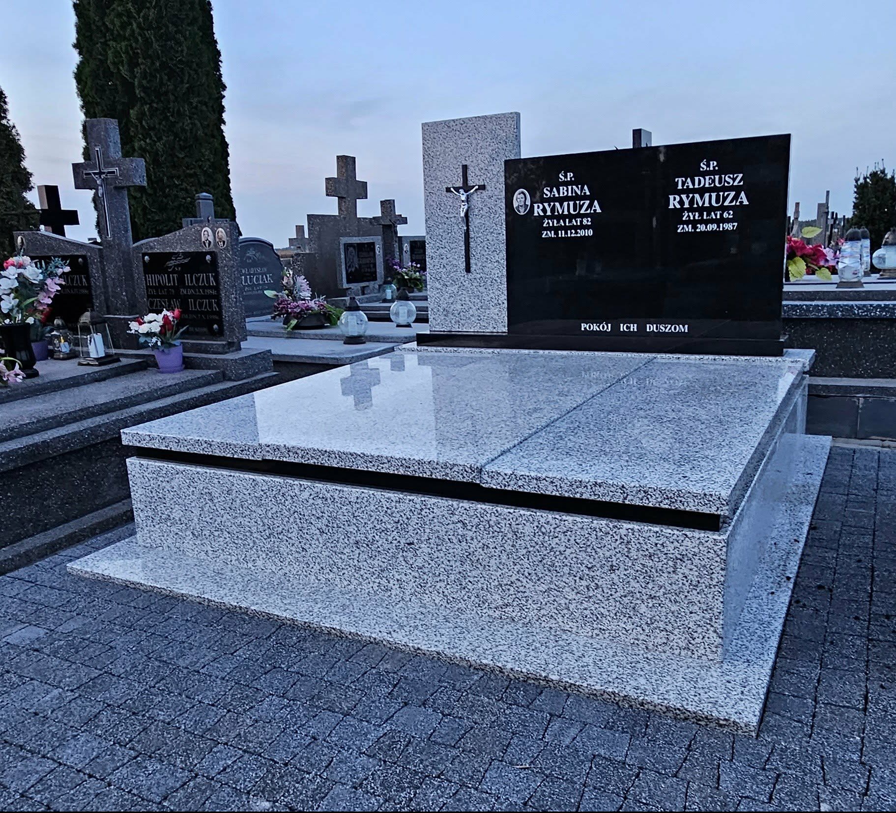 Granitowy nagrobek z czarną tablicą inskrypcyjną na cmentarzu, w tle inne groby i krzyże. Polerowana powierzchnia odbija niebo.