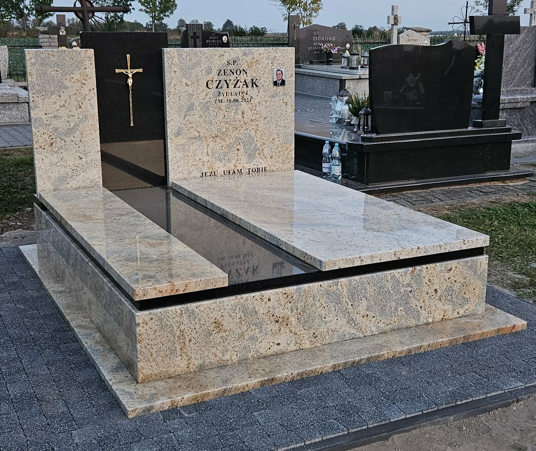 Jasny, granitowy nagrobek z czarnym elementem i złotym krzyżem, umieszczony na szarej kostce brukowej. Widoczne inskrypcje i zdjęcie osoby zmarłej.