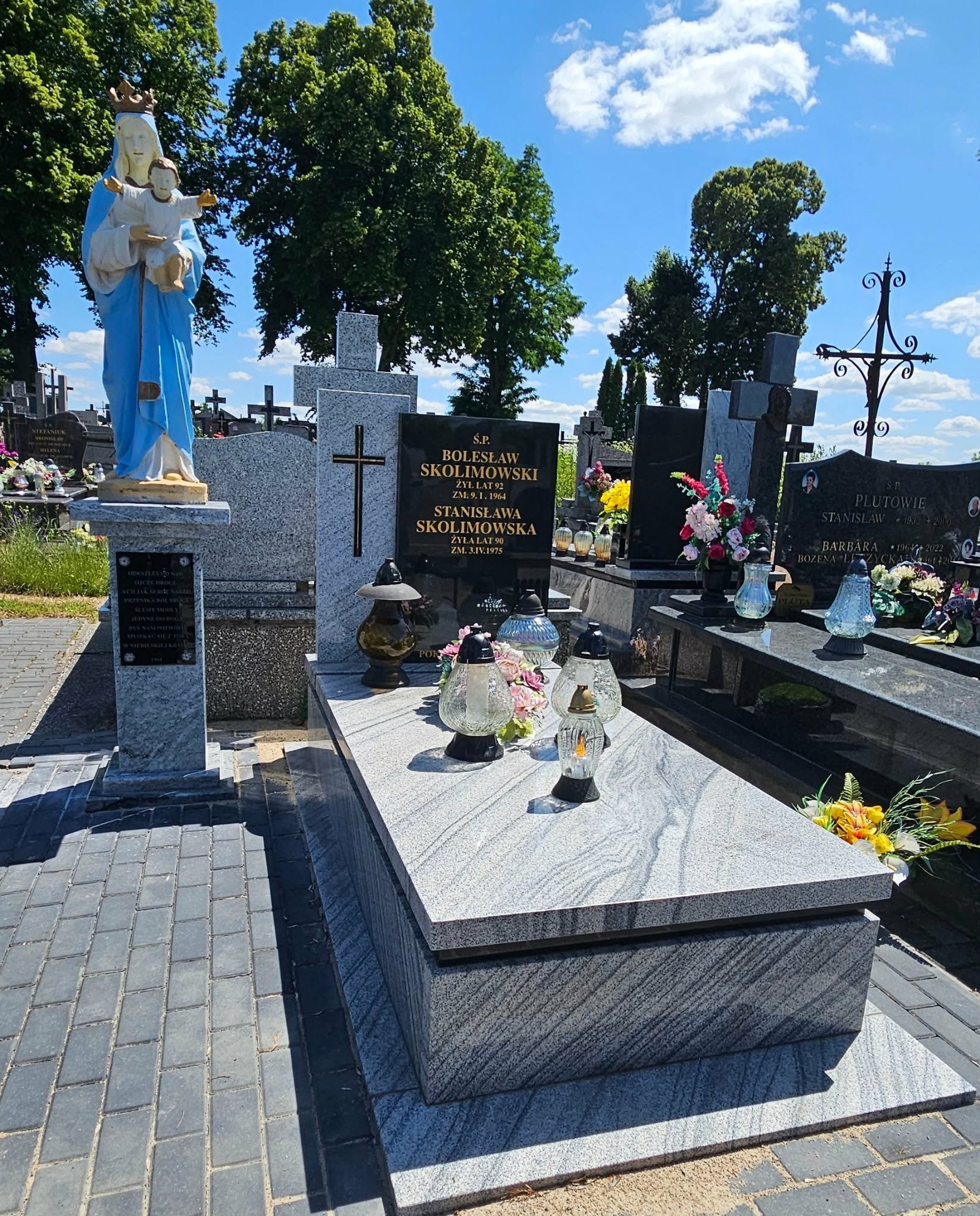 Granitowy nagrobek z jasnoszarą płytą, zdobiony lampionami i kwiatami, na cmentarzu w słoneczny dzień. W tle inne nagrobki i drzewa.