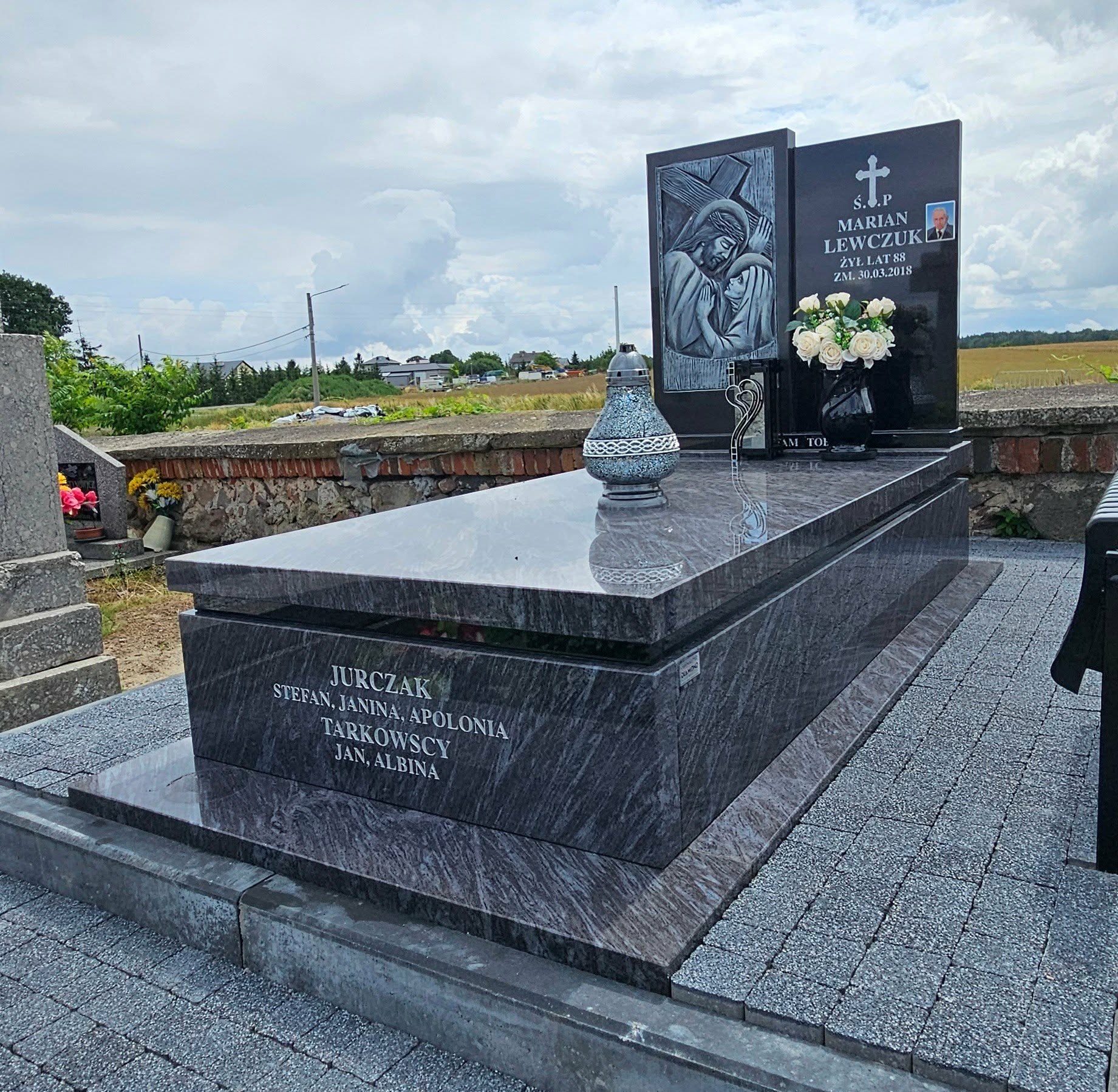 Granitowy nagrobek z inskrypcjami i wizerunkiem religijnym, otoczony kostką brukową. Na nagrobku wazon z białymi różami i lampion. Tło: krajobraz wiejski.