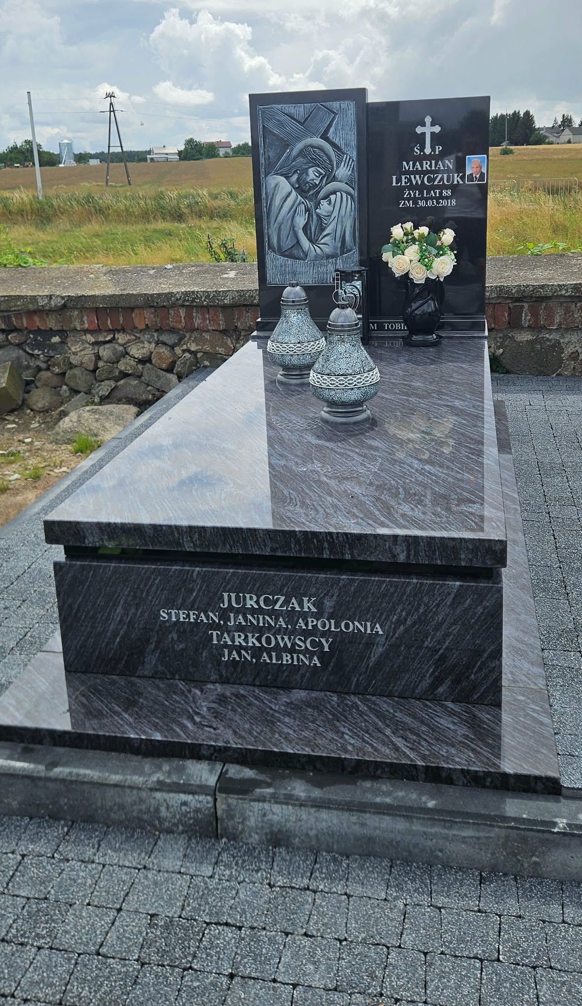 Granitowy nagrobek z inskrypcją nazwisk, zdobiony płaskorzeźbą i kwiatami, ustawiony na cmentarzu. W tle mur z kamienia i zielone pola.