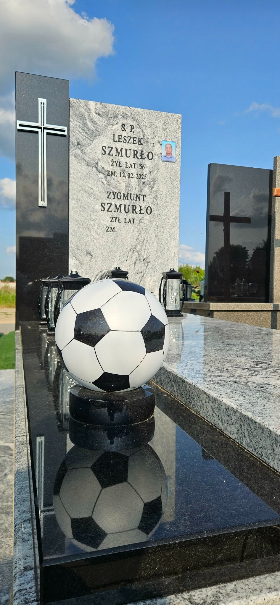 Granitowy nagrobek z piłką nożną, upamiętniający Leszka Szmurło. Widoczne lampiony i krzyż. Błękitne niebo w tle. Detal kamieniarski, refleksy na polerowanej powierzchni.