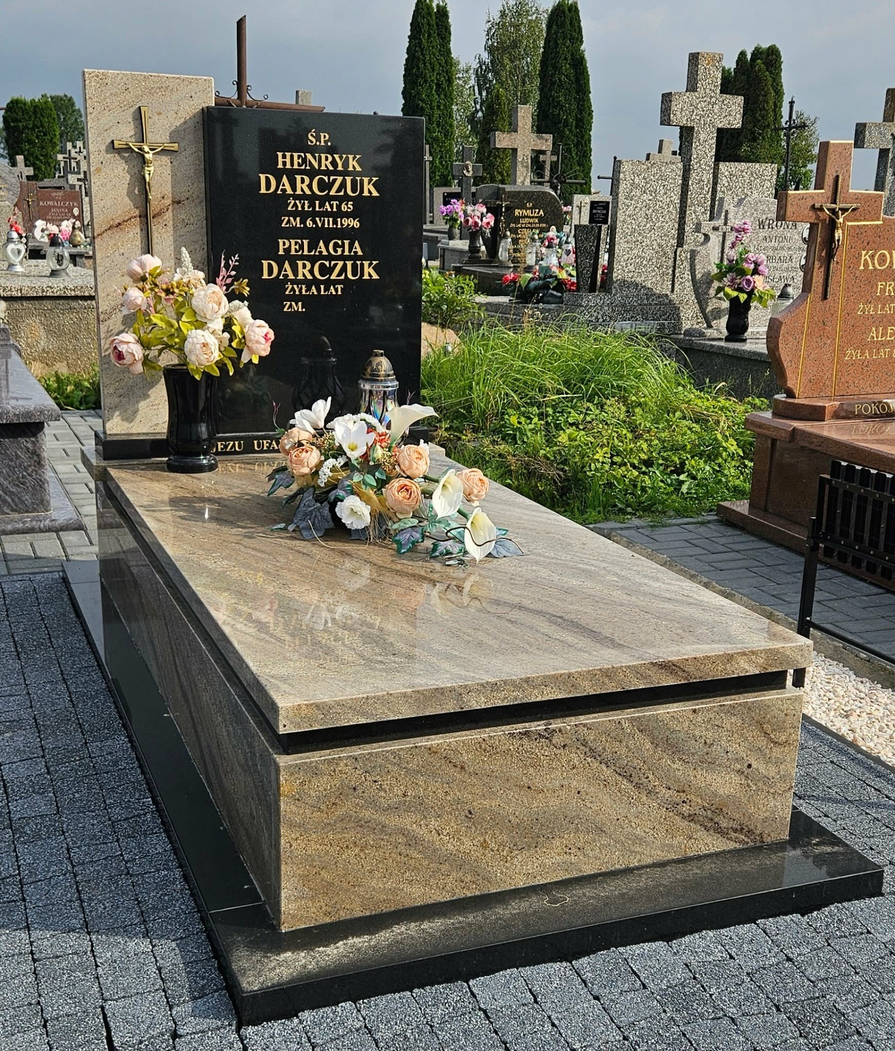Granitowy nagrobek z czarną tablicą inskrypcyjną i kwiatami, na cmentarzu. Jasny kamień nagrobka kontrastuje z ciemną kostką brukową. Widok z bliska, w tle inne nagrobki.