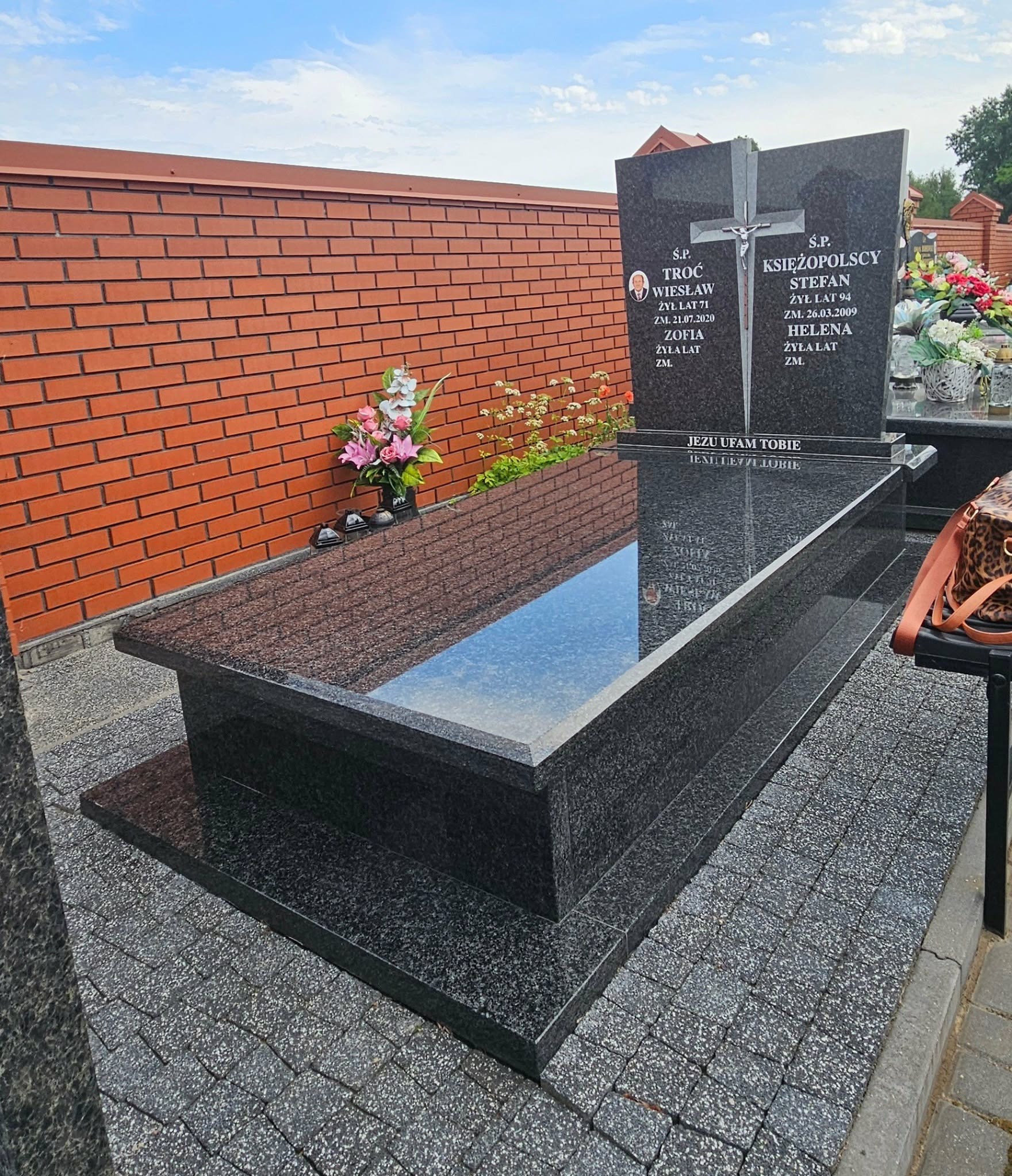 Granitowy nagrobek z krzyżem i inskrypcjami, otoczony kwiatami, na tle ceglanego muru. Polerowana powierzchnia odbija niebo. Całość na kostce brukowej.