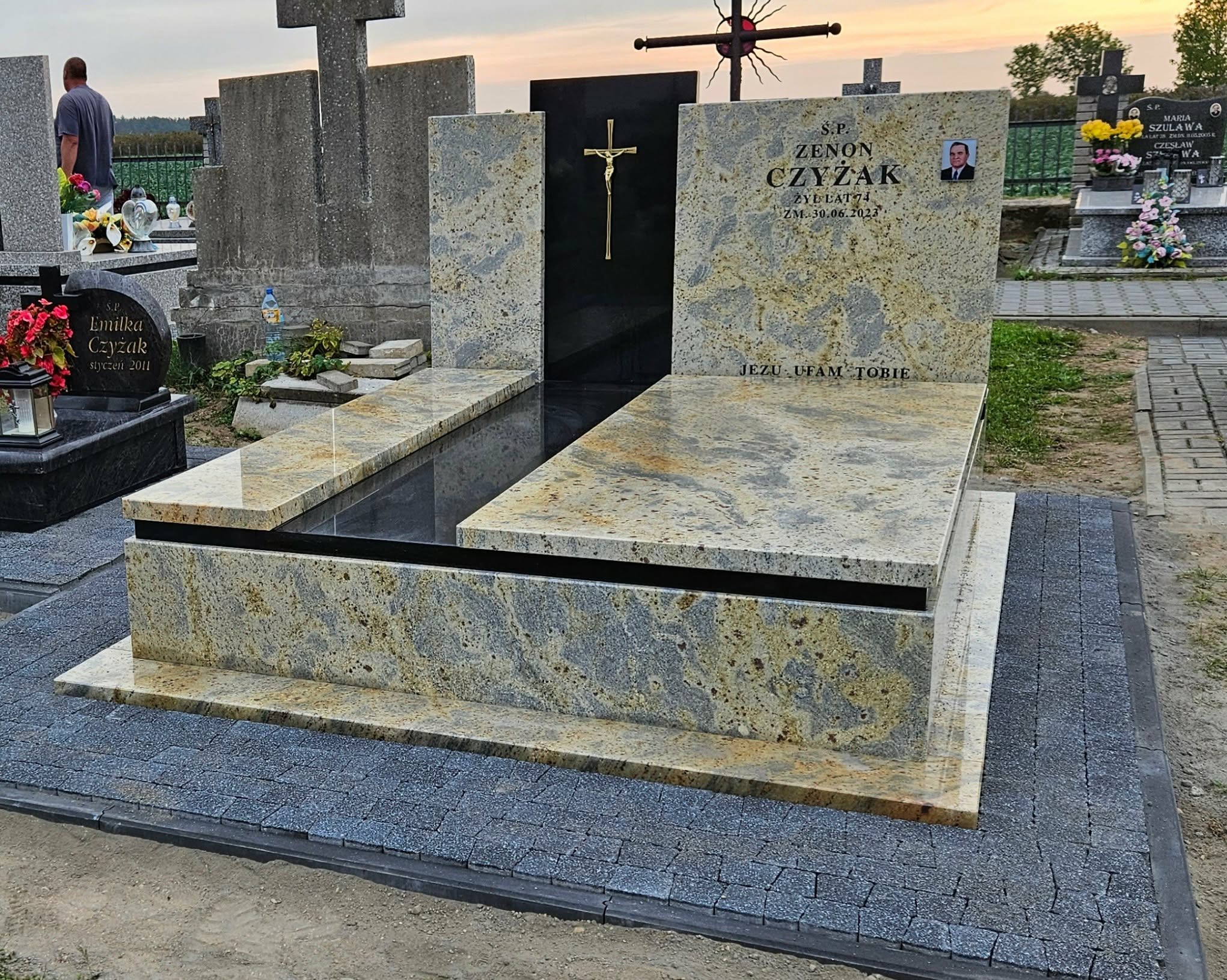 Granitowy nagrobek z czarnym elementem centralnym i złotym krucyfiksem, otoczony kostką brukową. Widoczne inskrypcje i zdjęcie zmarłego. Cmentarz w tle.