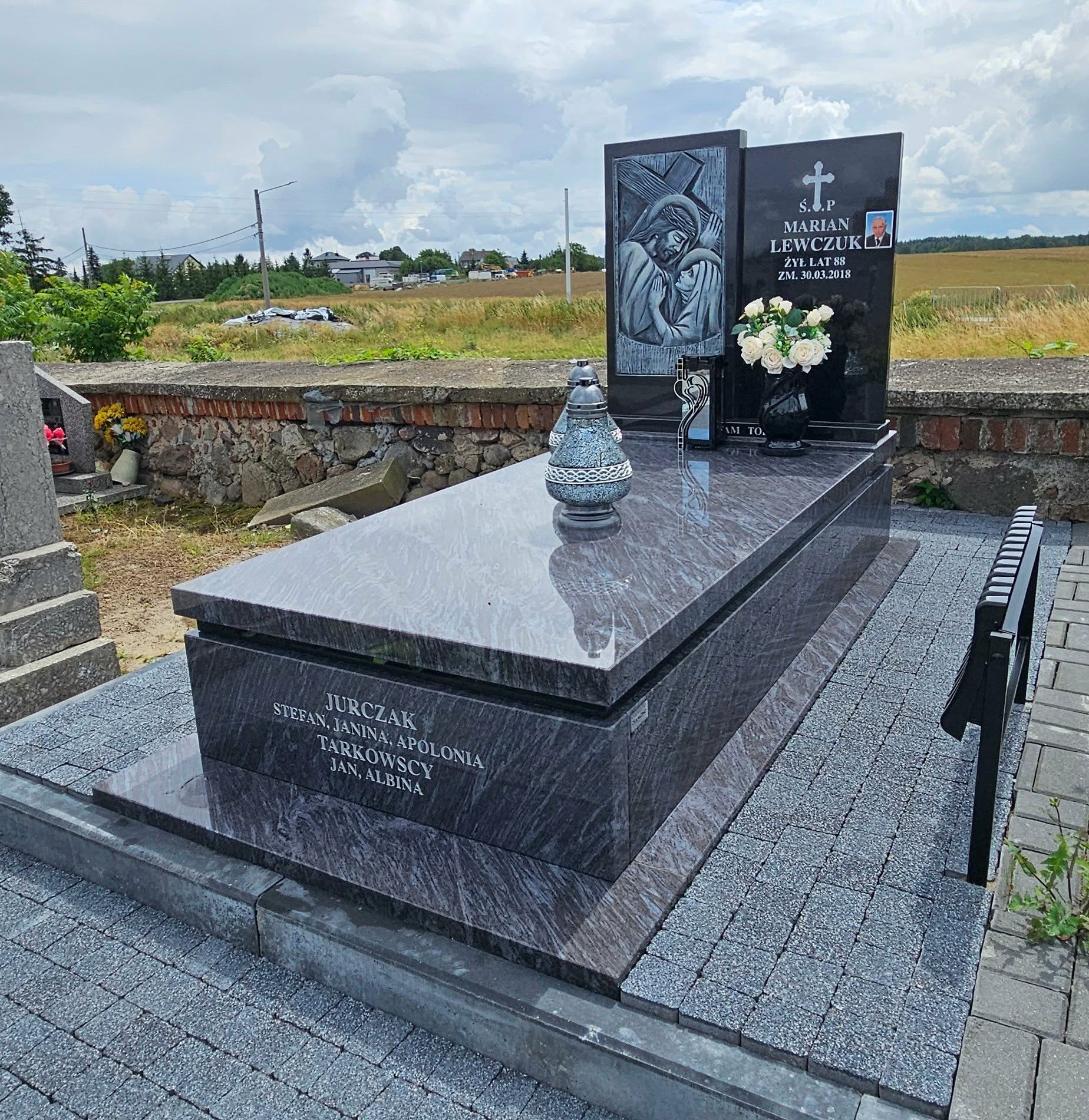 Granitowy nagrobek z inskrypcją i wizerunkiem religijnym, otoczony kostką brukową. Na płycie nagrobnej wazon z białymi kwiatami i lampion. Tło stanowi krajobraz polny.