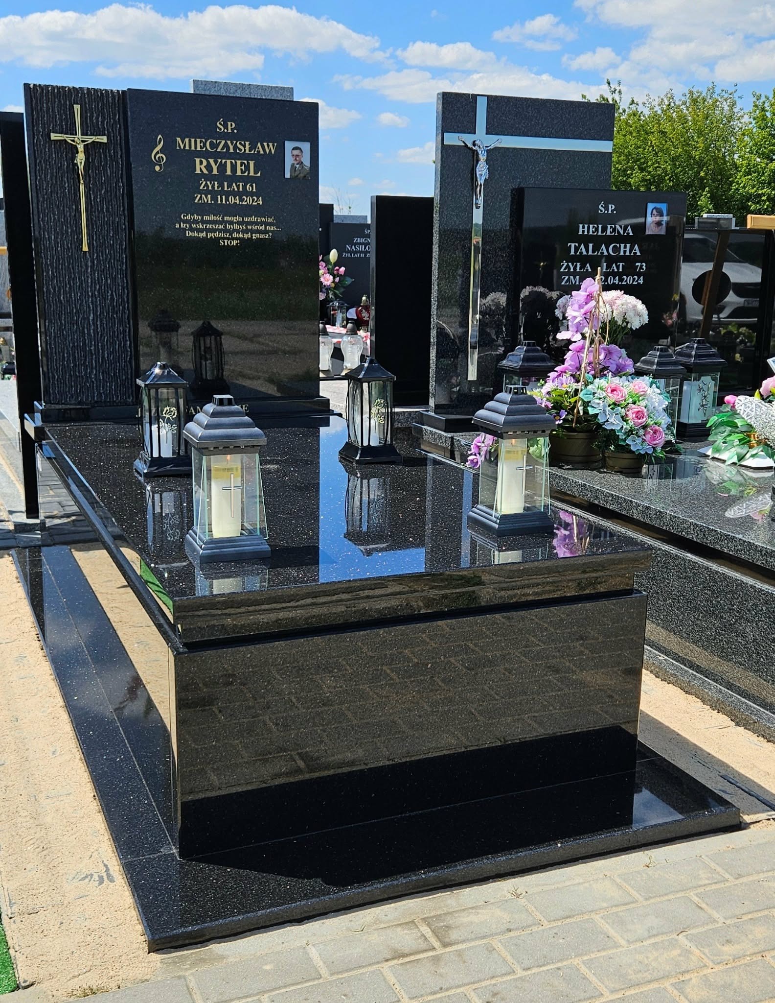 Czarny, polerowany nagrobek z granitu z lampionami i inskrypcjami, na tle innych nagrobków i zieleni cmentarza, w słoneczny dzień. Widoczne detale wykończenia.