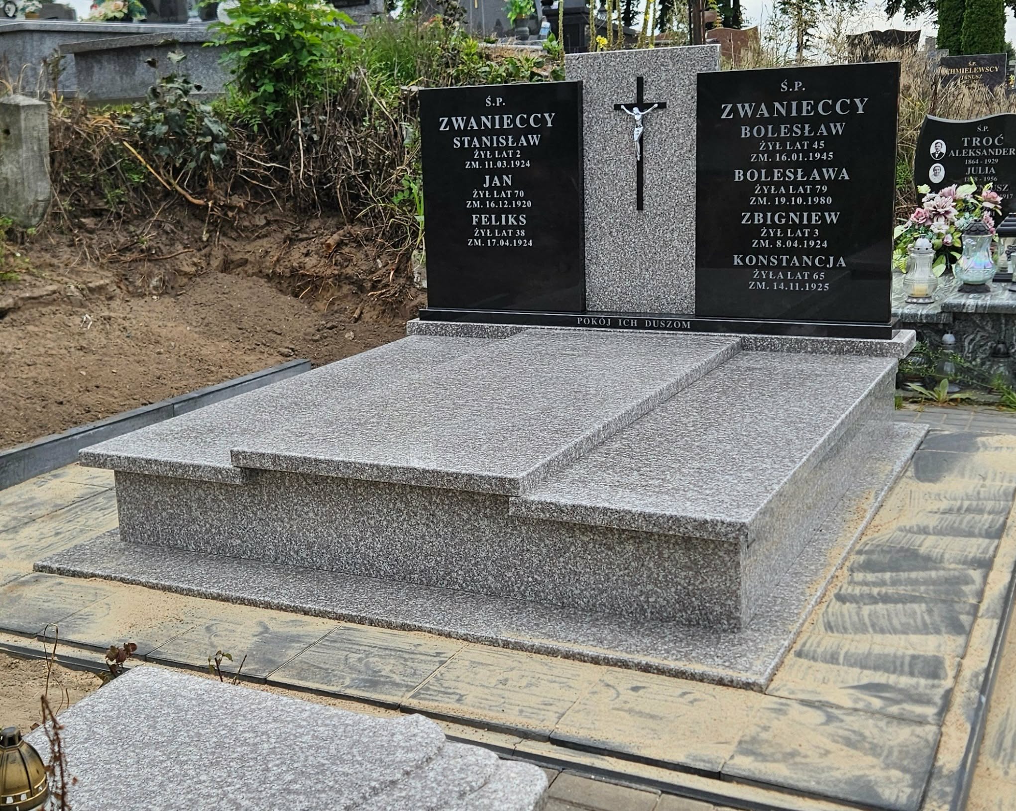 Granitowy nagrobek z czarnymi tablicami inskrypcyjnymi i krzyżem, otoczony zielenią cmentarza. Widoczne detale obróbki kamienia i inskrypcje.