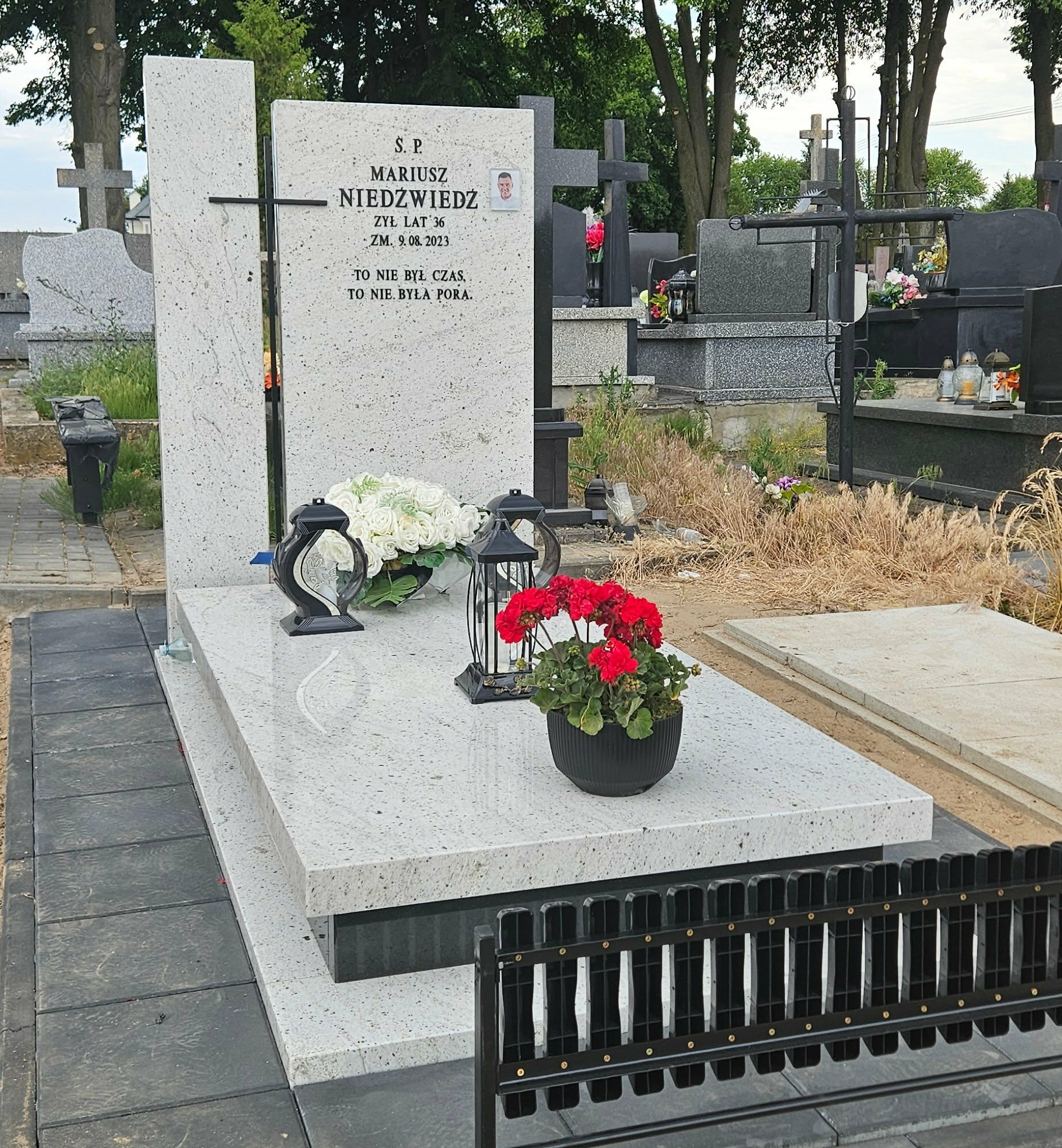 Jasny, granitowy nagrobek z czarnym krzyżem i zdjęciem osoby zmarłej, otoczony kwiatami i lampionami na cmentarzu. Widoczne inne nagrobki w tle.