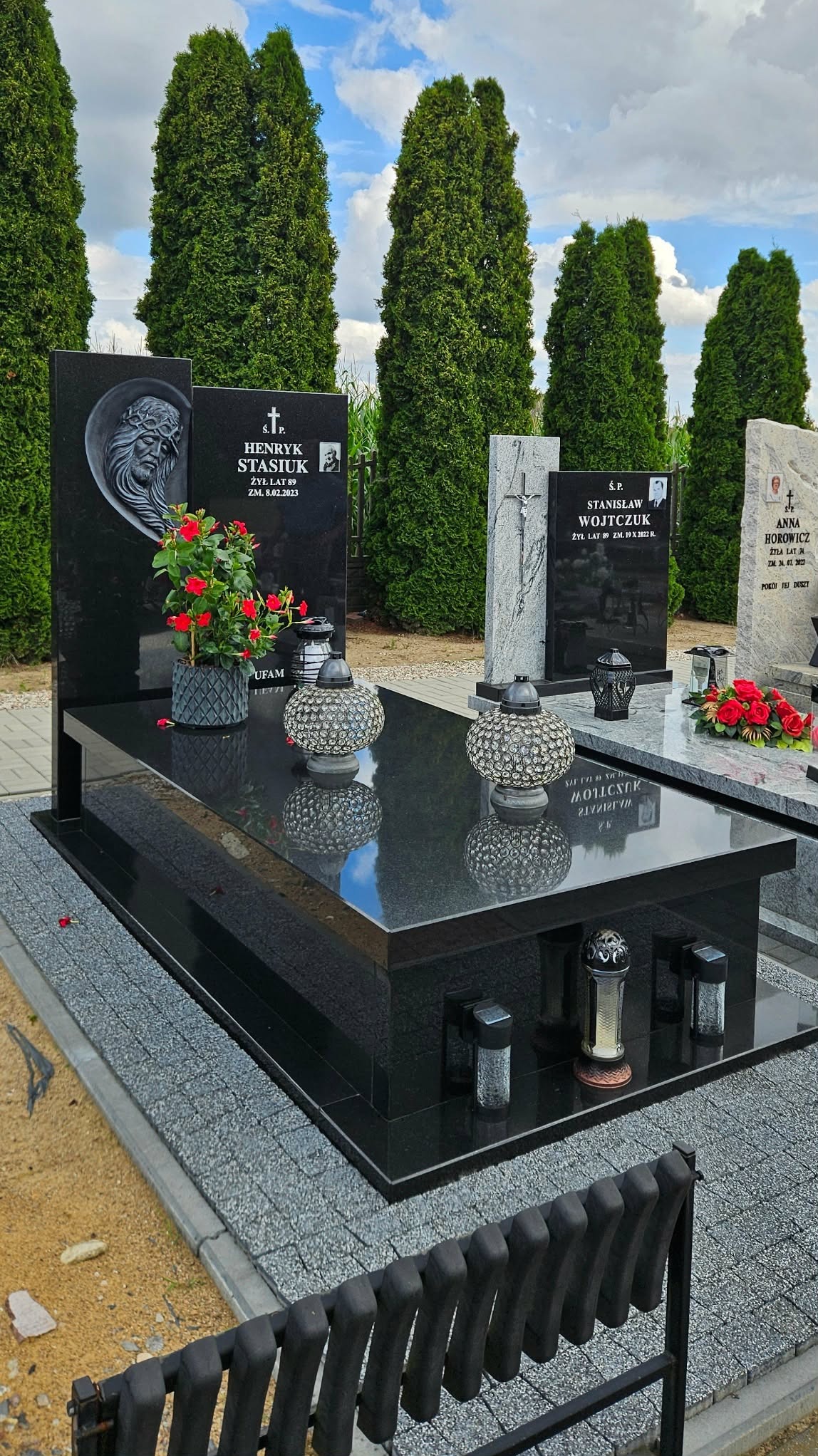 Czarny, podwójny nagrobek z granitu z kwiatami i lampionami, otoczony zielenią drzew. Widoczne inskrypcje i zdjęcia zmarłych. Całość na tle błękitnego nieba.