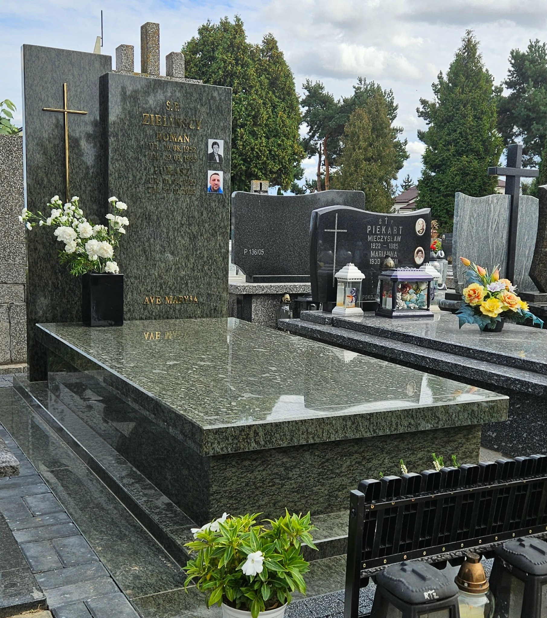 Granitowy nagrobek z inskrypcjami i zdjęciami, ozdobiony kwiatami w wazonie. Widoczne inne nagrobki w tle, cmentarz w pochmurny dzień.