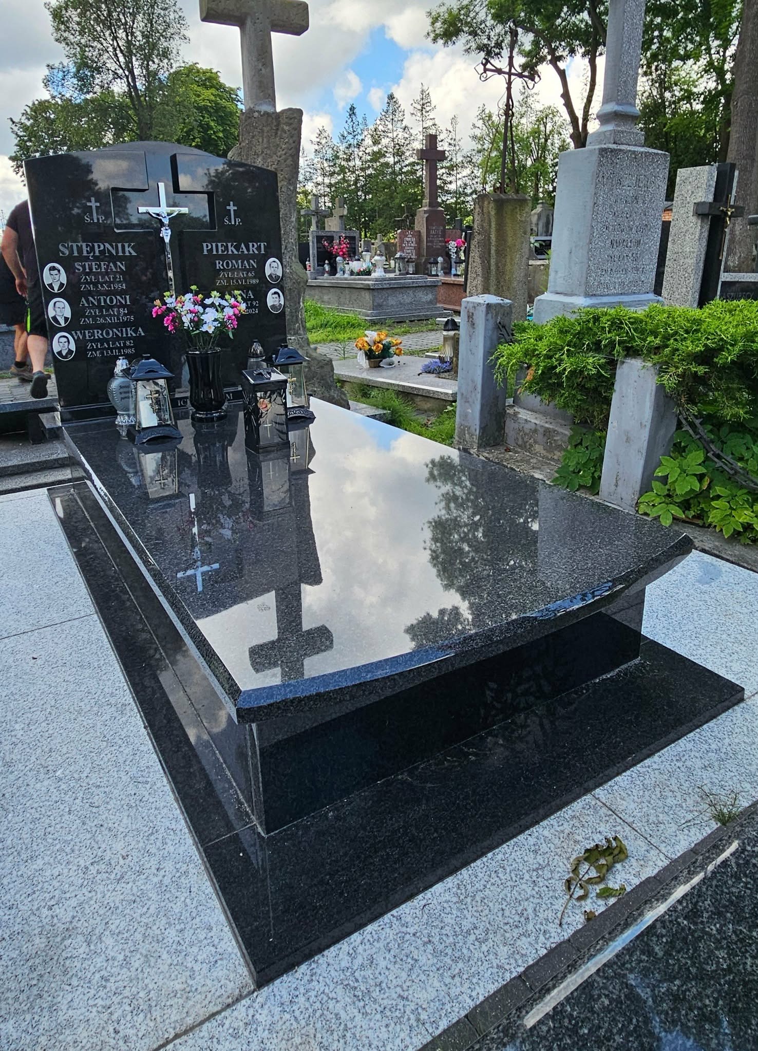 Czarny, polerowany nagrobek z inskrypcjami i zdjęciami zmarłych, ozdobiony kwiatami i lampionami, na cmentarzu z innymi nagrobkami w tle.