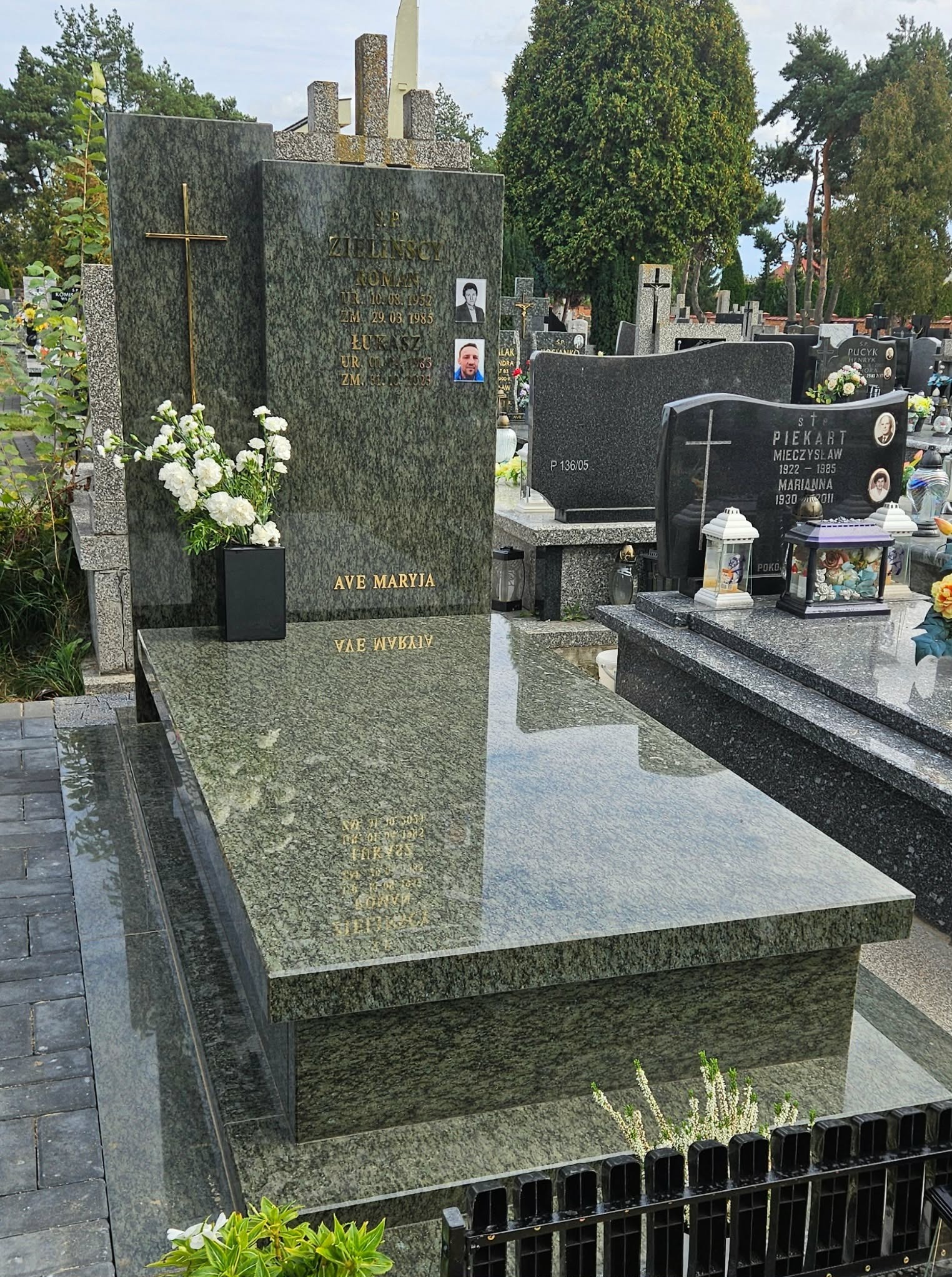 Granitowy nagrobek z krzyżem i zdjęciami zmarłych, otoczony innymi nagrobkami na cmentarzu. Na płycie nagrobnej napis 'AVE MARYJA' i wyryte dane zmarłych.