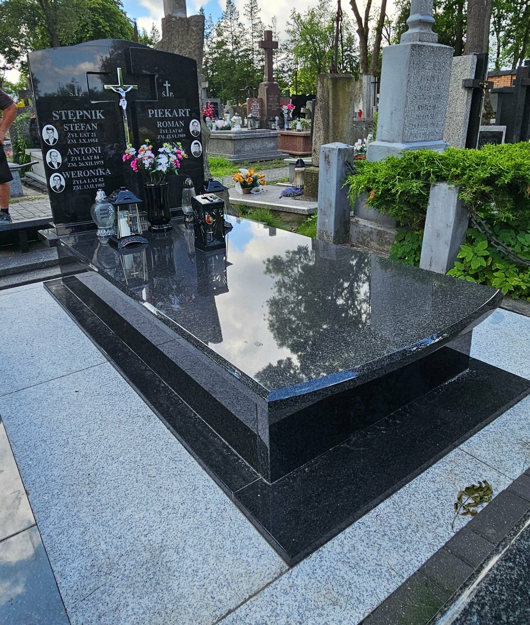 Czarny, polerowany nagrobek z granitu na cmentarzu, z wygrawerowanymi danymi zmarłych i kwiatami. W tle inne nagrobki i drzewa. Widoczne odbicie nieba w polerowanej powierzchni.