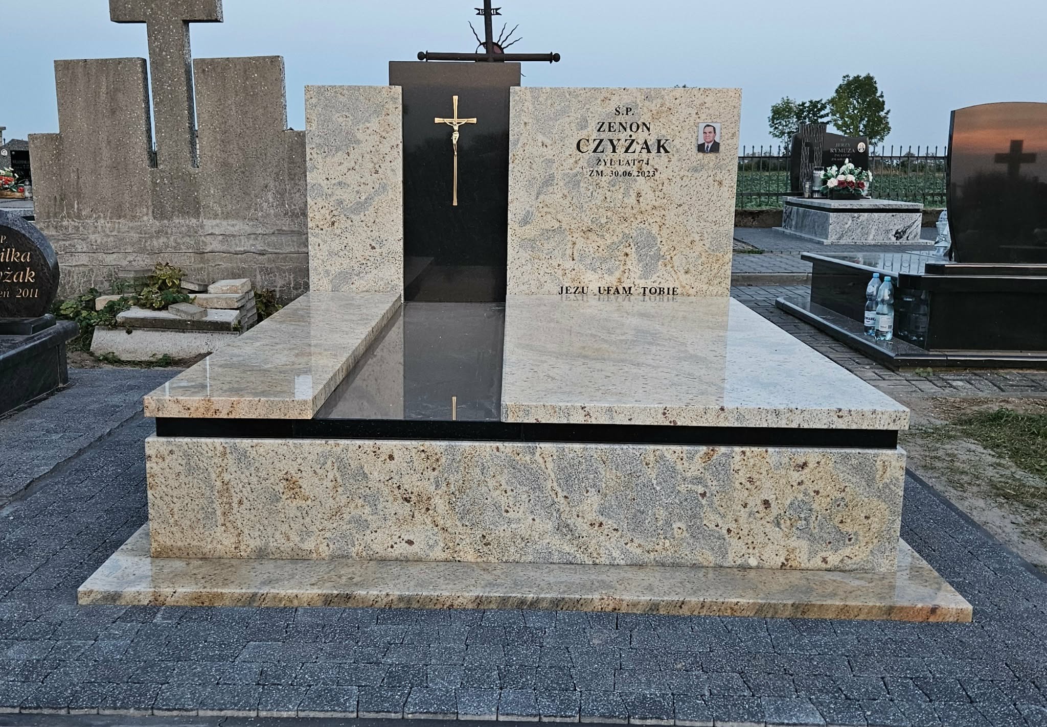 Granitowy nagrobek z krzyżem i portretem osoby zmarłej, umieszczony na cmentarzu. Jasny kamień z ciemnymi elementami, otoczony kostką brukową. Widok z przodu, w tle inne nagrobki.