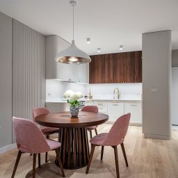 Pracownia mebli Cami - Nowoczesna kuchnia z drewnianymi szafkami, okrągłym stołem i różowymi krzesłami. Białe blaty i oświetlenie LED podkreślają minimalistyczny design.