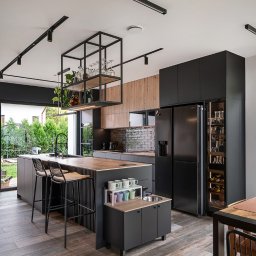 Pracownia mebli Cami - Nowoczesna kuchnia z wyspą i czarnymi szafkami, drewniane blaty, oświetlenie szynowe, przeszklona lodówka na wino, widok na ogród przez okno.