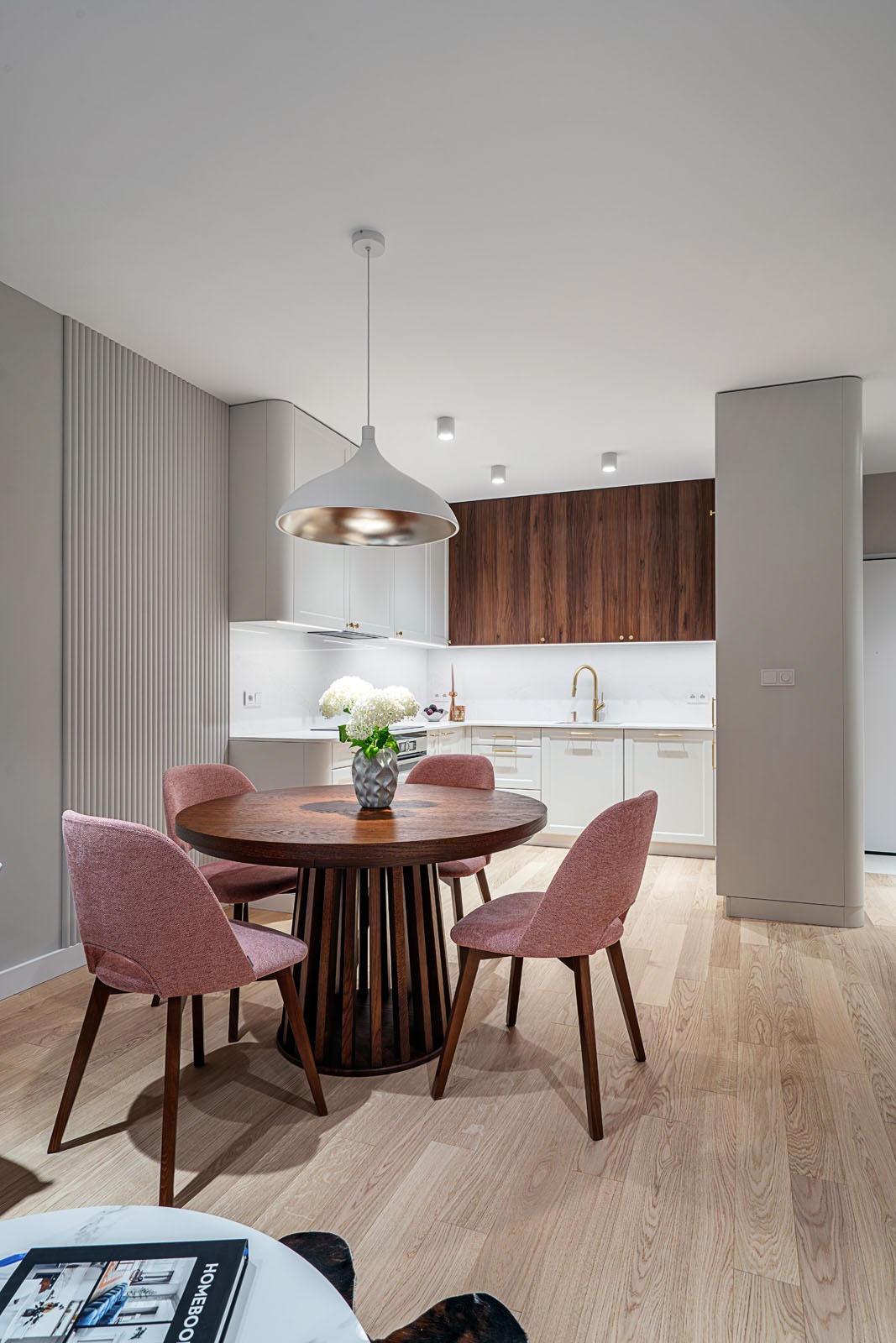 Nowoczesna kuchnia z drewnianymi szafkami, okrągłym stołem i różowymi krzesłami. Białe blaty i oświetlenie LED podkreślają minimalistyczny design.