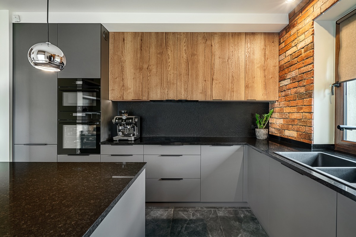 Szara kuchnia z drewnianymi szafkami wiszącymi i ceglaną ścianą. Na blacie ekspres do kawy i roślina doniczkowa. Nad wyspą kuchenną wisi chromowana lampa.
