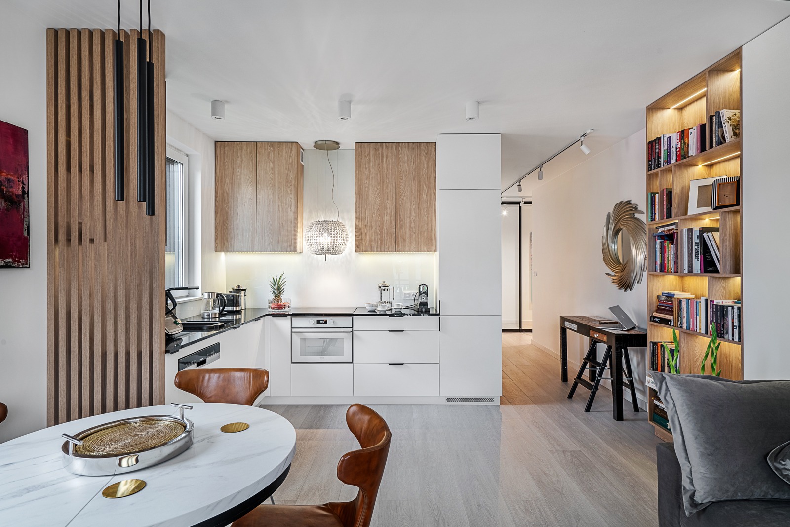 Nowoczesna kuchnia z białymi szafkami i drewnianymi akcentami, okrągły stół z marmurowym blatem i skórzanymi krzesłami, oświetlona półka na książki w tle.