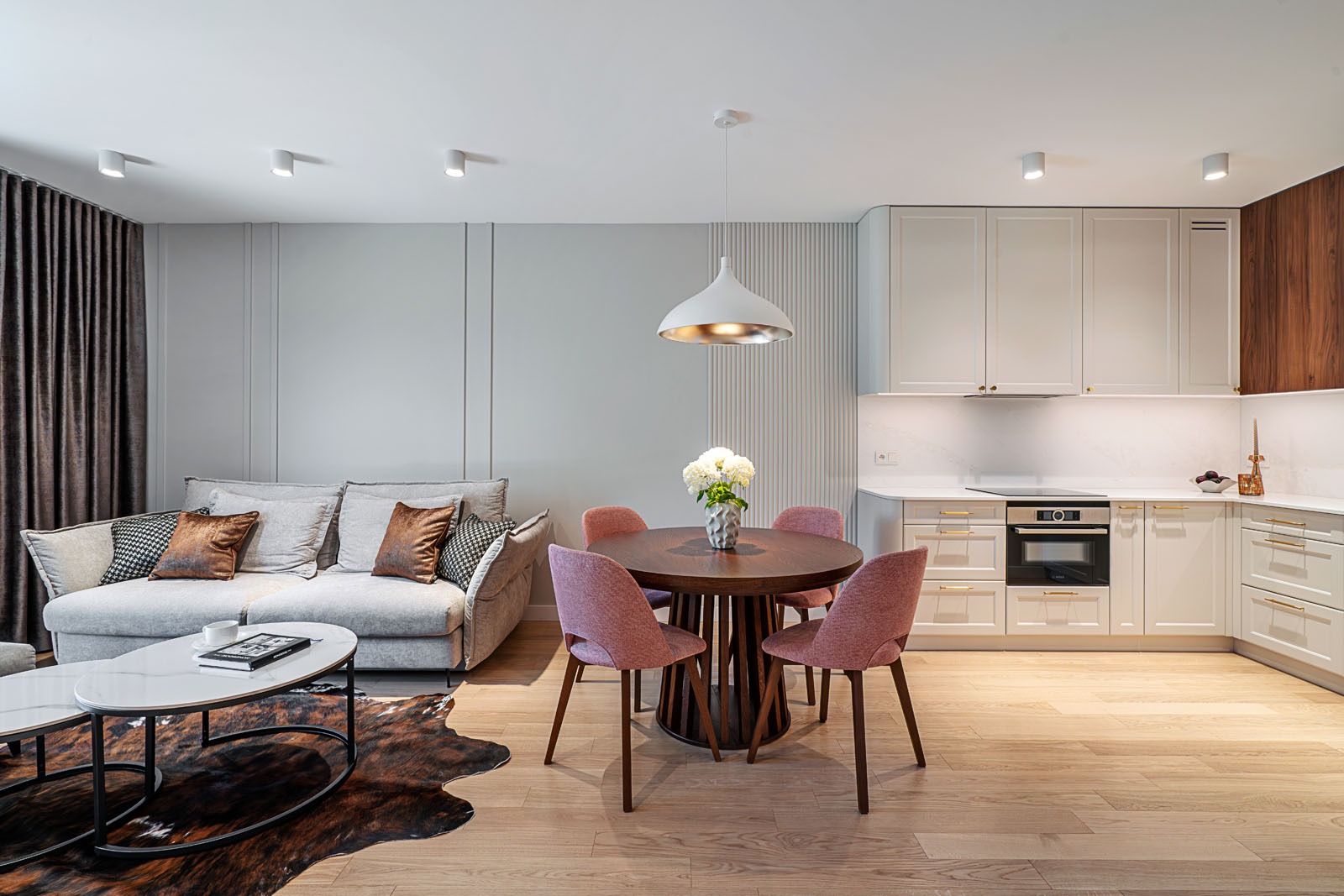 Jasne, nowoczesne wnętrze z aneksem kuchennym i okrągłym stołem w Lanckoronie. Kremowe szafki kuchenne, drewniany blat, różowe krzesła i szara sofa tworzą spójną całość.