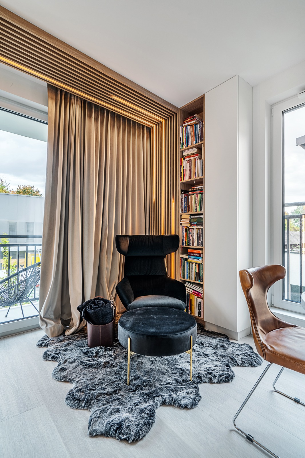Stylowy kącik czytelniczy z czarnym fotelem i pufą na futrzanym dywanie, obok regał z książkami i okno z widokiem na balkon. Drewniane lamele z oświetleniem.