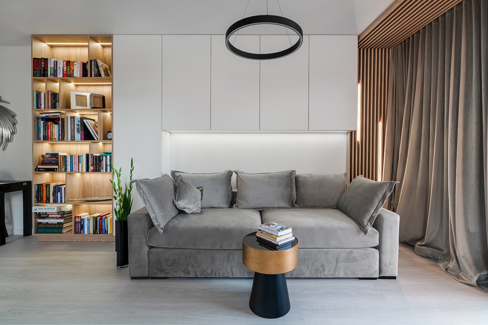 Stylowy salon z szarą sofą, drewnianą biblioteczką z podświetleniem LED i minimalistycznym oświetleniem. Elementy dekoracyjne i zasłony w odcieniach beżu.