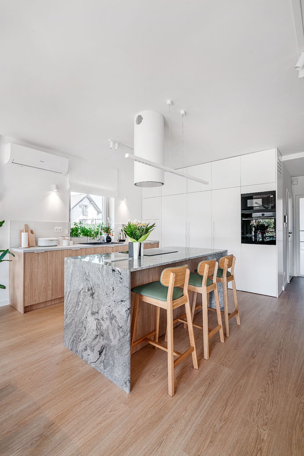 Jasna kuchnia z wyspą o marmurowym blacie i drewnianymi hokerami. Minimalistyczny design, białe szafki i drewniane akcenty. Nowoczesne oświetlenie.