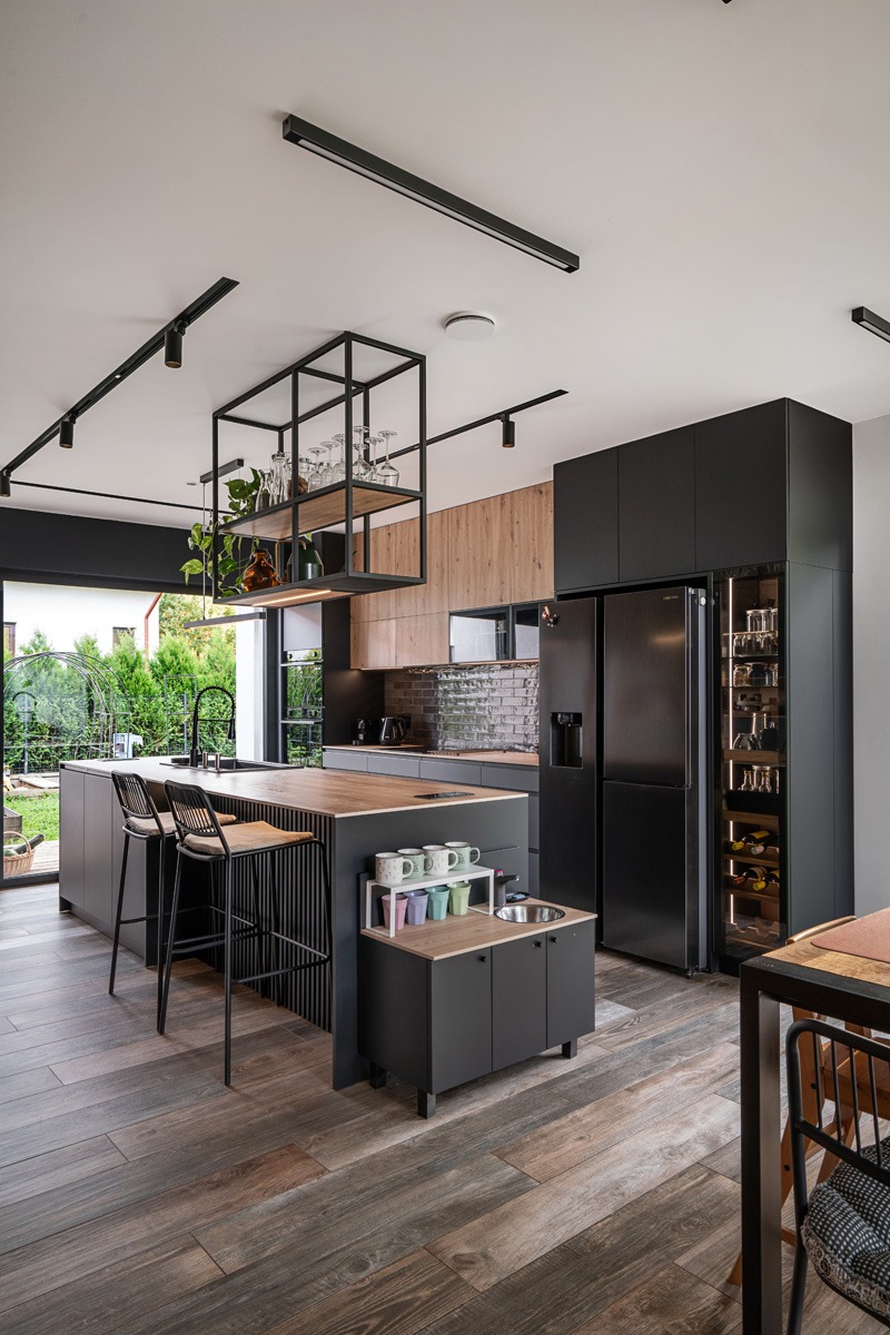 Nowoczesna kuchnia z wyspą i czarnymi szafkami, drewniane blaty, oświetlenie szynowe, przeszklona lodówka na wino, widok na ogród przez okno.