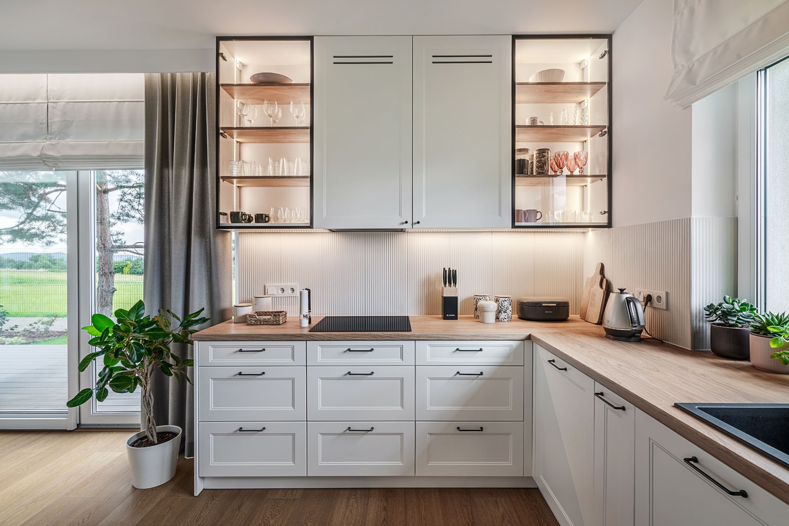 Jasna kuchnia z drewnianym blatem i przeszklonymi szafkami podświetlonymi LED, z widokiem na zieleń za oknem. Minimalistyczny design z rośliną doniczkową.