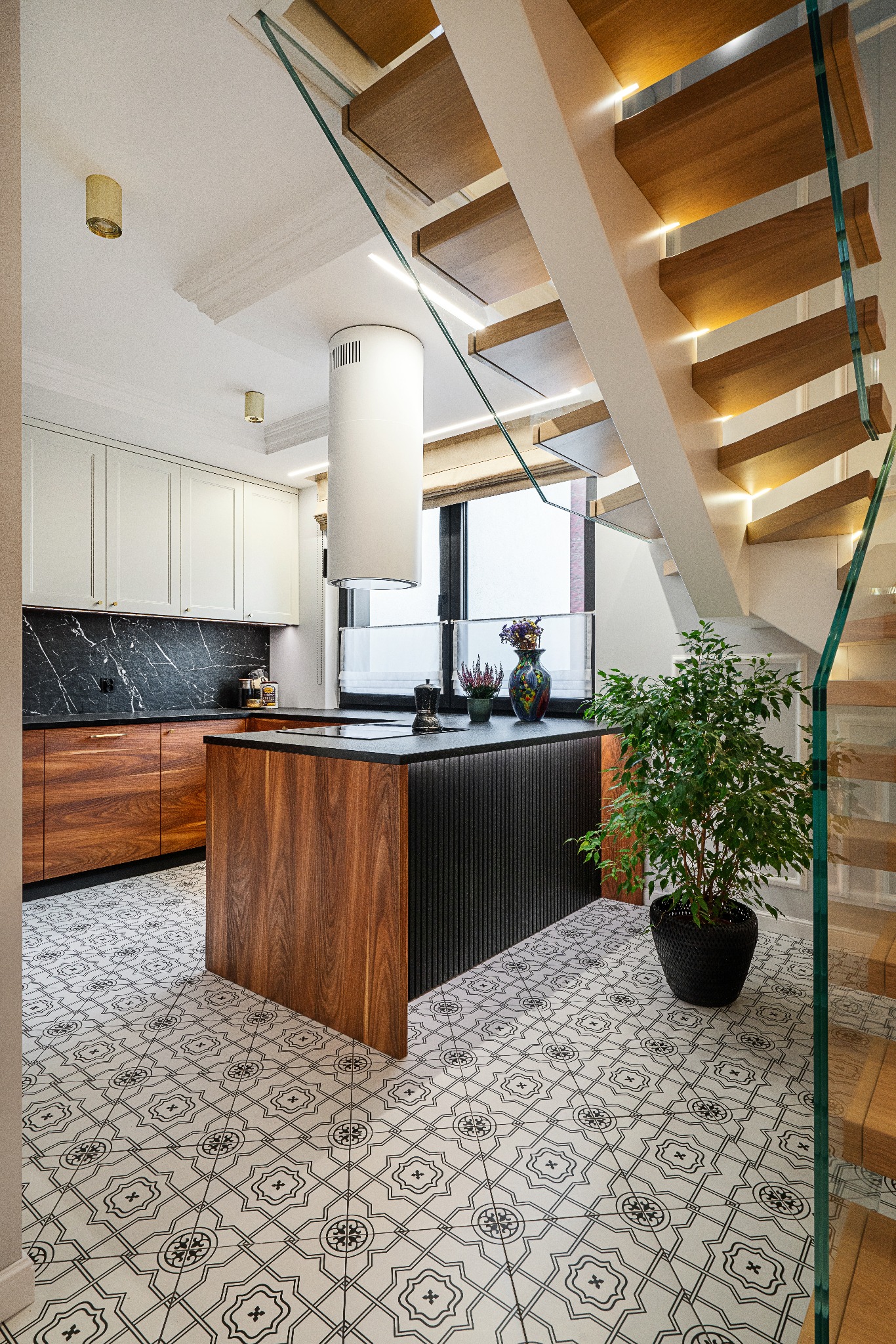 Nowoczesna kuchnia z wyspą, drewnianymi frontami i czarnym blatem, połączona ze schodami z drewnianymi stopniami i szklaną balustradą. W tle okno z kwiatami.