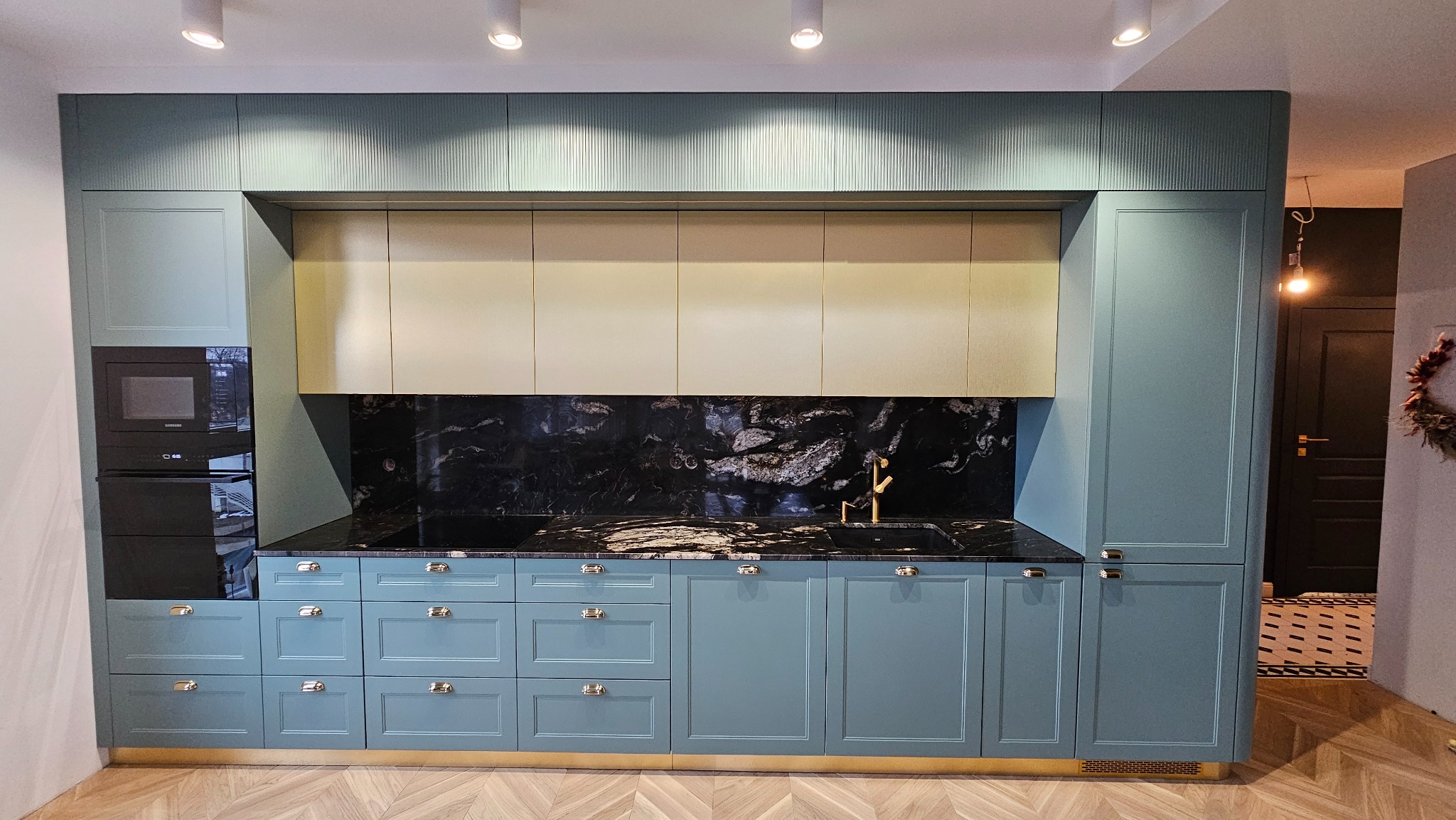 Niebieska kuchnia na wymiar z czarnym blatem i złotą armaturą. Górne szafki w kolorze kremowym. Podłoga z parkietu ułożonego w jodełkę. Widok na fragment przedpokoju.