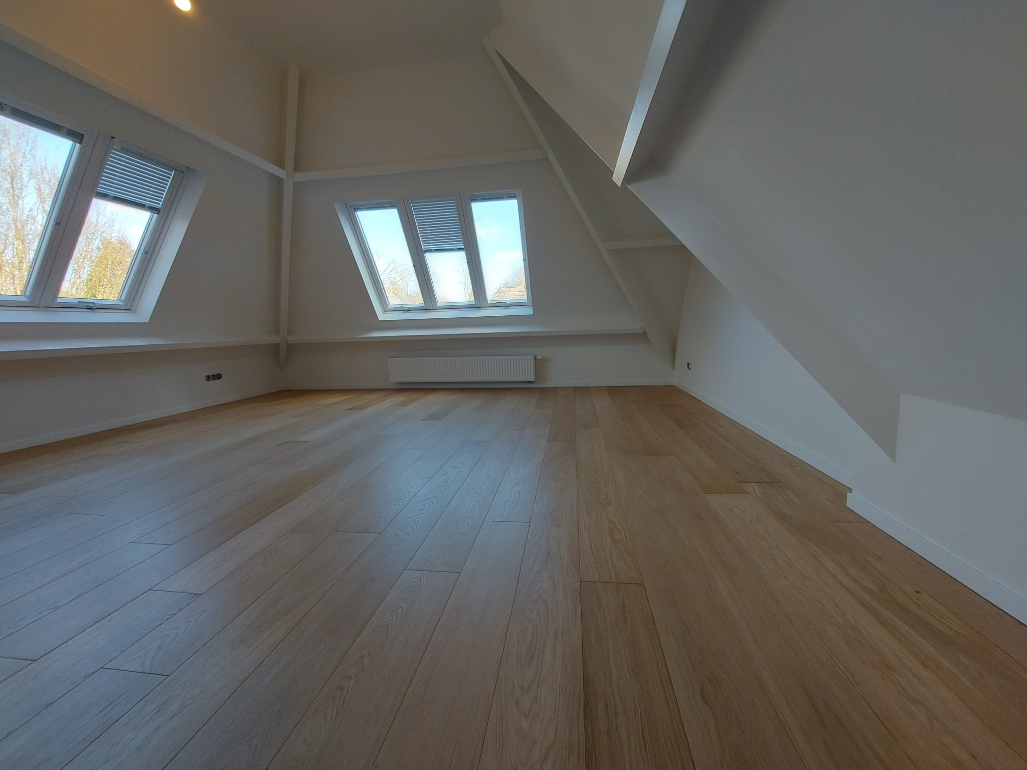 Jasne, przestronne poddasze po remoncie. Parkiet z naturalnego drewna, białe ściany i okna dachowe z żaluzjami. Minimalistyczny design, skosy i nowoczesny grzejnik.