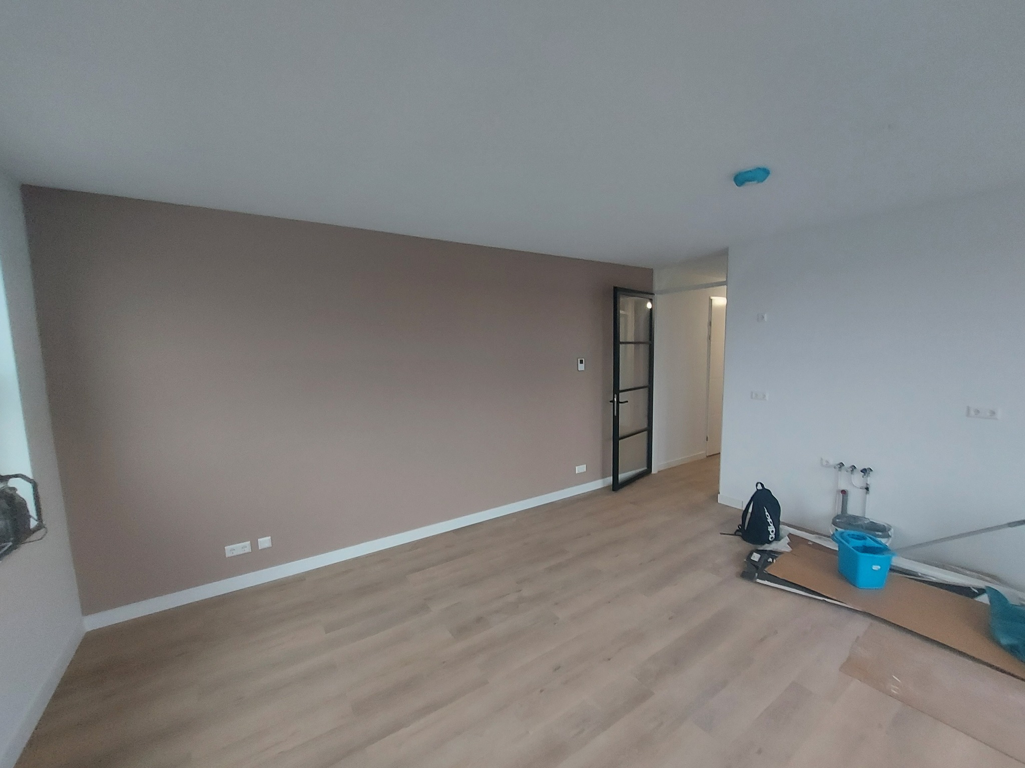 Pomieszczenie po malowaniu: beżowa ściana kontrastuje z białą, drewniana podłoga, widoczne narzędzia malarskie i czarny plecak. Drzwi z czarną ramą w tle.