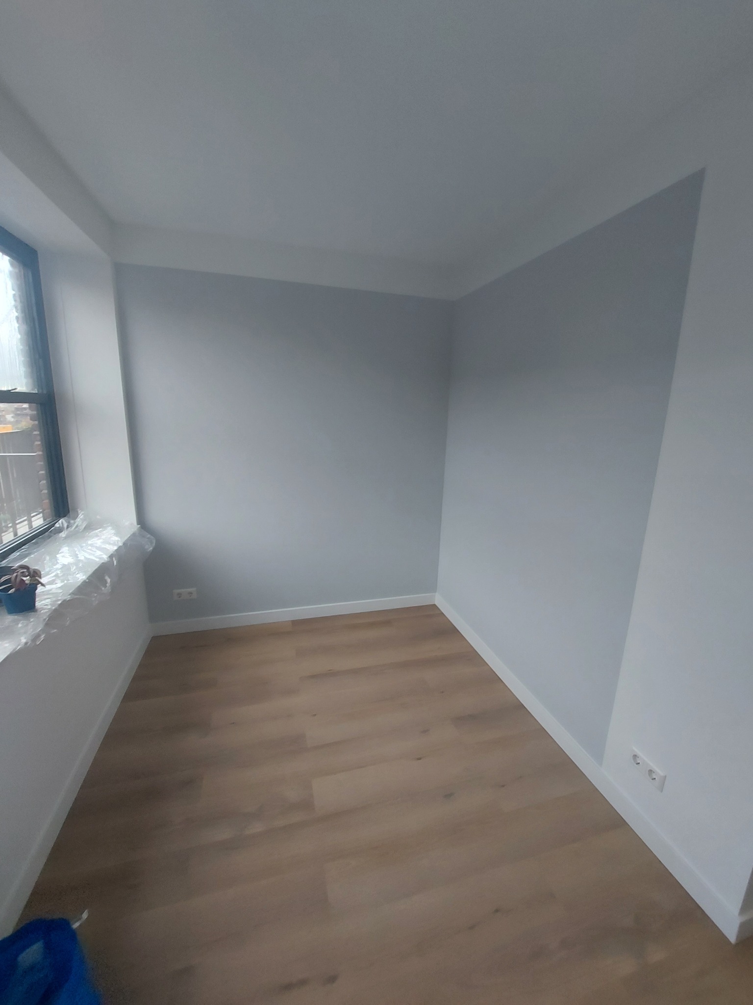 Wnętrze pokoju po malowaniu ścian na szaro, z drewnianą podłogą i białymi listwami przypodłogowymi. Okno zabezpieczone folią malarską.