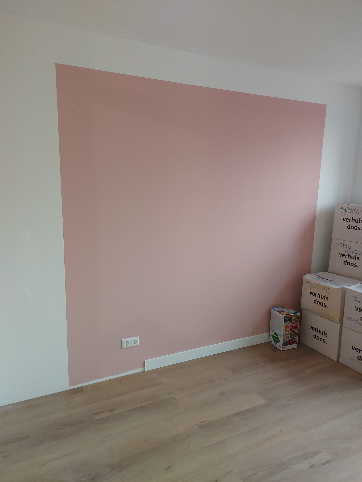 Ściana pomalowana na różowo w pokoju z drewnianą podłogą. Widoczne białe listwy przypodłogowe i gniazdka elektryczne. Kartony z napisem 'verhuis doos' w rogu.