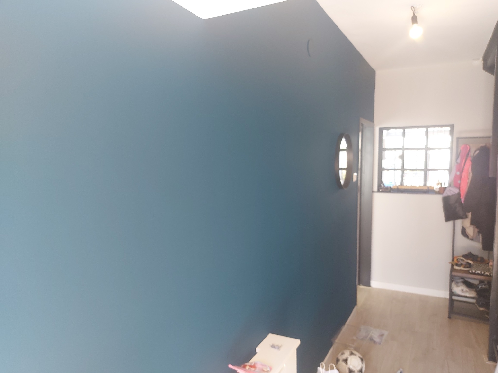 Ściana pomalowana na niebiesko w przedpokoju z oknem w kratkę i okrągłym lustrem. Widoczny fragment szafki na buty i piłka. Świeże, matowe wykończenie.