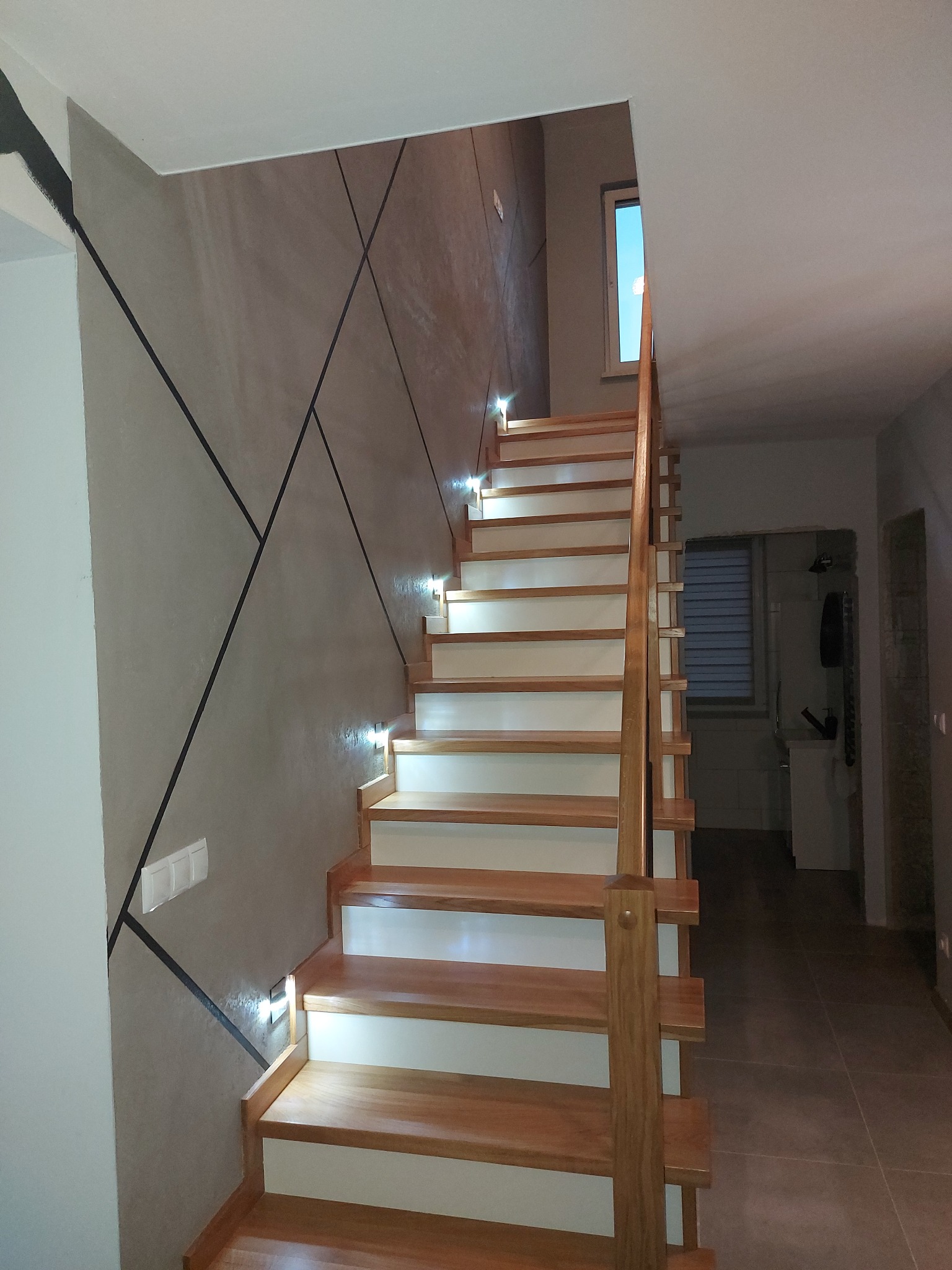 Nowoczesne schody drewniane z oświetleniem LED wkomponowane w ścianę z geometrycznym wzorem. Widoczne detale wykończenia i estetyczne połączenie materiałów.