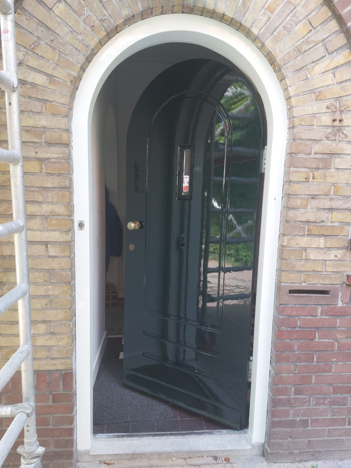 Błyszczące, ciemnozielone drzwi wejściowe, częściowo otwarte, w łukowatym, ceglanym portalu z białym obramowaniem. Widoczny fragment rusztowania.